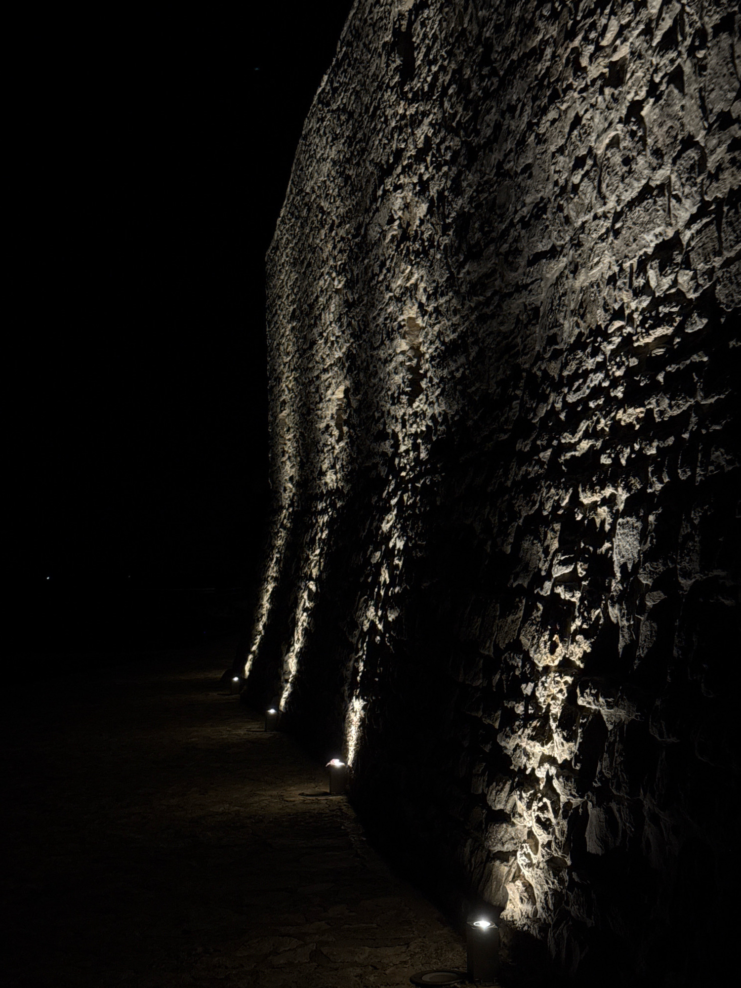 The image depicts a pathway illuminated by low-lying lights at the base of a rough, textured stone wall. The wall is uneven, with natural formations and shadows cast by the upward lighting. The pathway is dark and appears to be made of earth or gravel. The lighting creates dramatic patterns on the wall, highlighting its rugged surface. The scene is set at night, with the background fading into complete darkness. There are no people visible in the image. This pathway and wall appear to be part of the historic fortifications of Novigrad, Croatia, a coastal town known for its well-preserved medieval architecture and defensive walls.