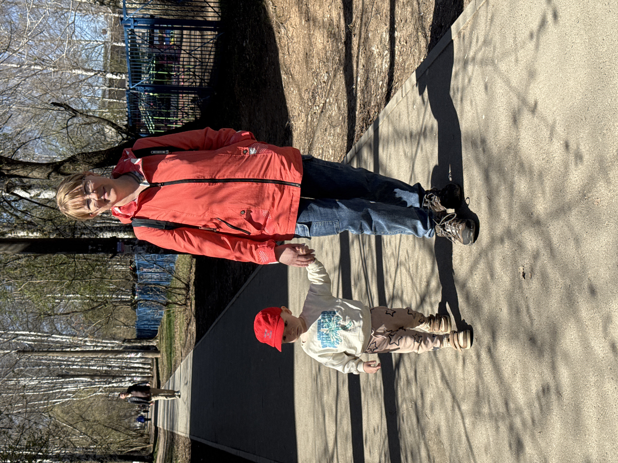 A woman and a young child are standing on a paved walkway outdoors. The woman is wearing glasses, a red jacket, and blue jeans. The child is wearing a red cap, a white sweatshirt, and patterned pants. They are holding hands. The walkway is lined with trees and a blue metal fence in the background. The location is Izhevsk, Russia.