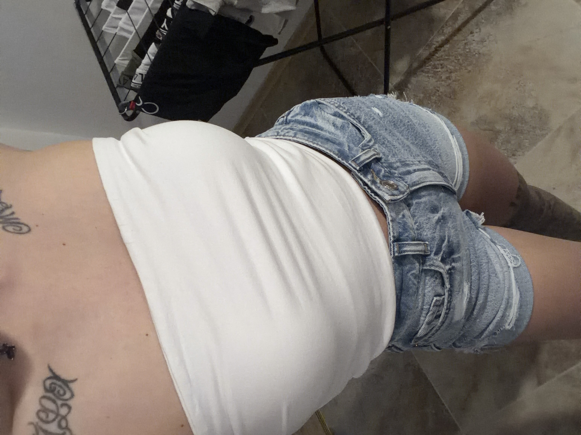 The image depicts an indoor scene featuring the torso and upper legs of a person from an overhead perspective. The person wears a white strapless tube top and light blue distressed denim shorts with frayed edges, visible pockets, and a button closure. Tattoos are present on the upper left chest area, featuring stylized black lettering, and on the upper right chest, showing a black swirling design. A dark-patterned tattoo is partially visible on the right thigh. The background on the right includes a light grey wall and a dark metal clothes drying rack holding various items of clothing, including black and white garments. The floor consists of large, light-toned tiles. The specific outdoor location in Plovdiv, Bulgaria, is not discernible from the visual information provided within the image, which solely shows an indoor setting. No direct actions or interactions are visible beyond the person standing for the photograph.