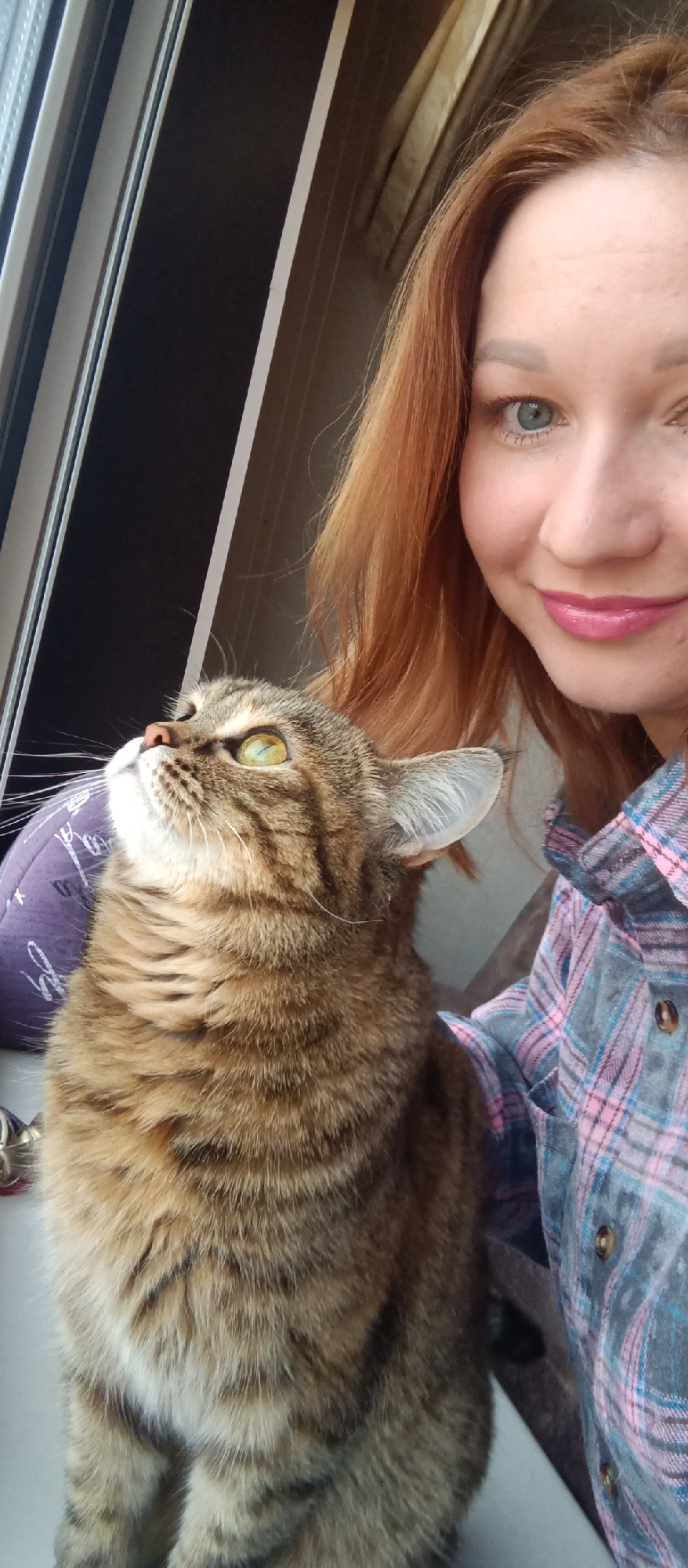 A woman and a cat are posing for a selfie. The woman has red hair, blue eyes, and is wearing a pink shirt. The cat is a tabby with yellow eyes and is looking up. They are next to a window. Location: Unknown, Unknown.