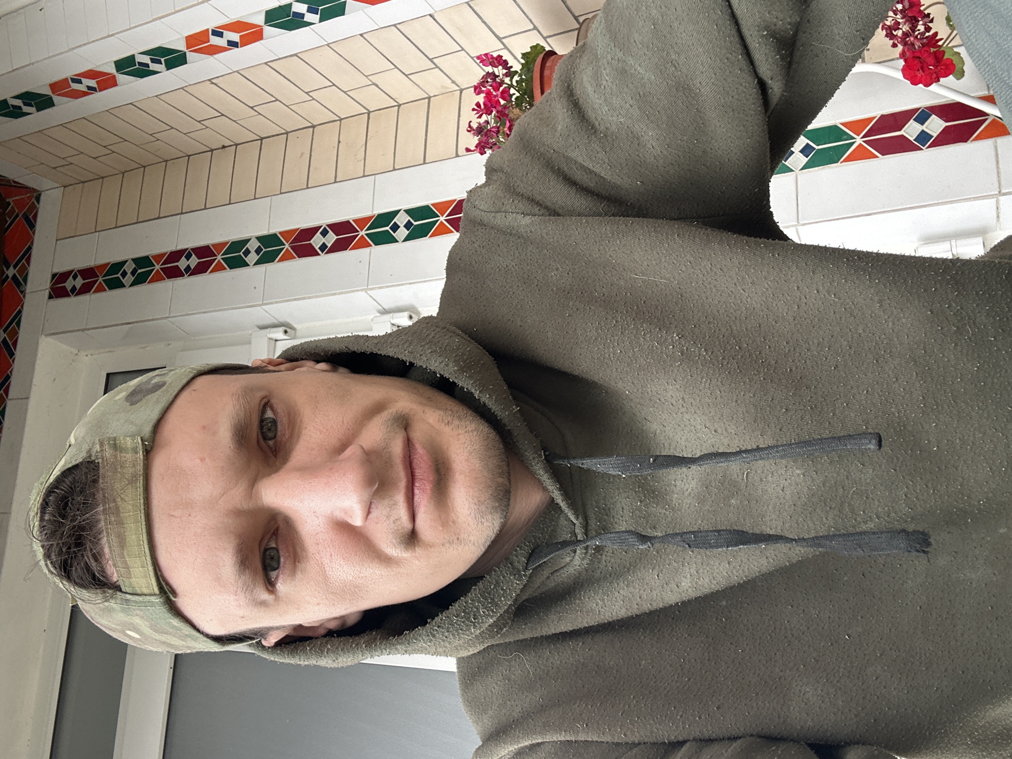 A man wearing a camouflage baseball cap and a dark green hoodie is taking a selfie. The man has light skin, a light brown stubble, and grey eyes. He is looking directly at the camera with a neutral expression. The hoodie appears worn, with small pills of fabric visible on its surface. The background shows a tiled wall with a geometric pattern of white, beige, green, orange, and red tiles. There is a potted plant with pink flowers on the right side of the image. The scene suggests an outdoor or semi-outdoor location, possibly a balcony or porch, in Makiv, Ukraine.