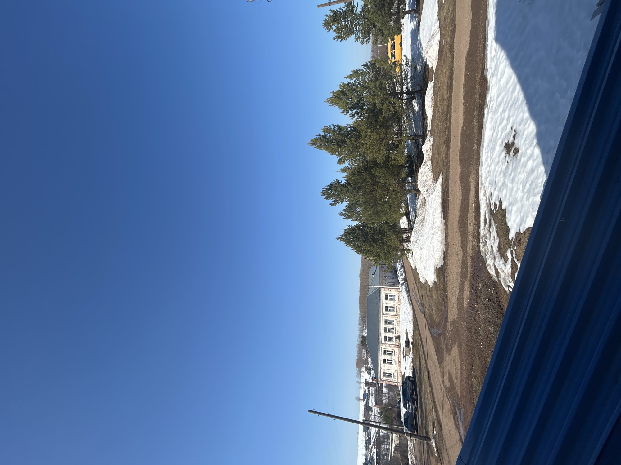 The image shows a landscape with a building in the distance. The sky is clear and blue. In the foreground, there are patches of snow and bare ground. Trees line the side of a dirt road. A yellow school bus is parked near some of the trees. A multi-story brick building with many windows can be seen in the distance. Some cars are parked near the building. There are telephone poles in the scene. The image appears to be taken from an elevated position. The blue metal roof is in the very foreground. The location is Фоки, Russia.
