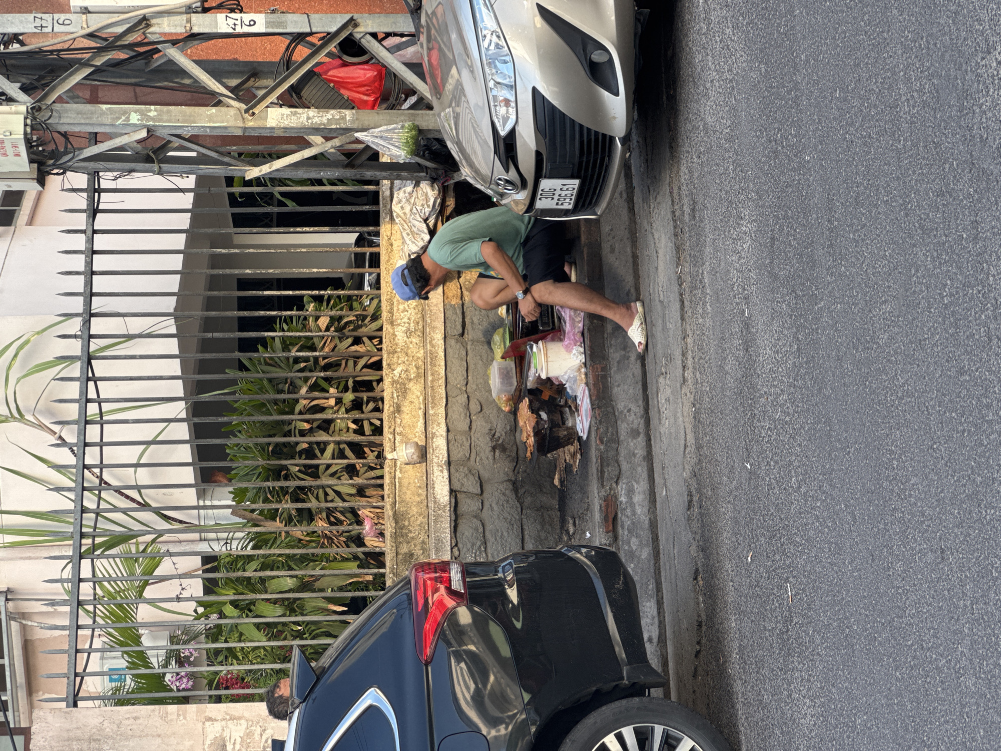 A street scene in Nha Trang Ward, Vietnam features a man in a green t-shirt, black shorts, and a blue baseball cap sitting on a curb beside a light-colored sedan. He is looking down at a small charcoal grill where food is being cooked. A black SUV is parked to his left. The background shows a building with a metal fence and lush vegetation. There are also electrical poles and wires visible. The ground is paved with asphalt and concrete, with some litter scattered around.
