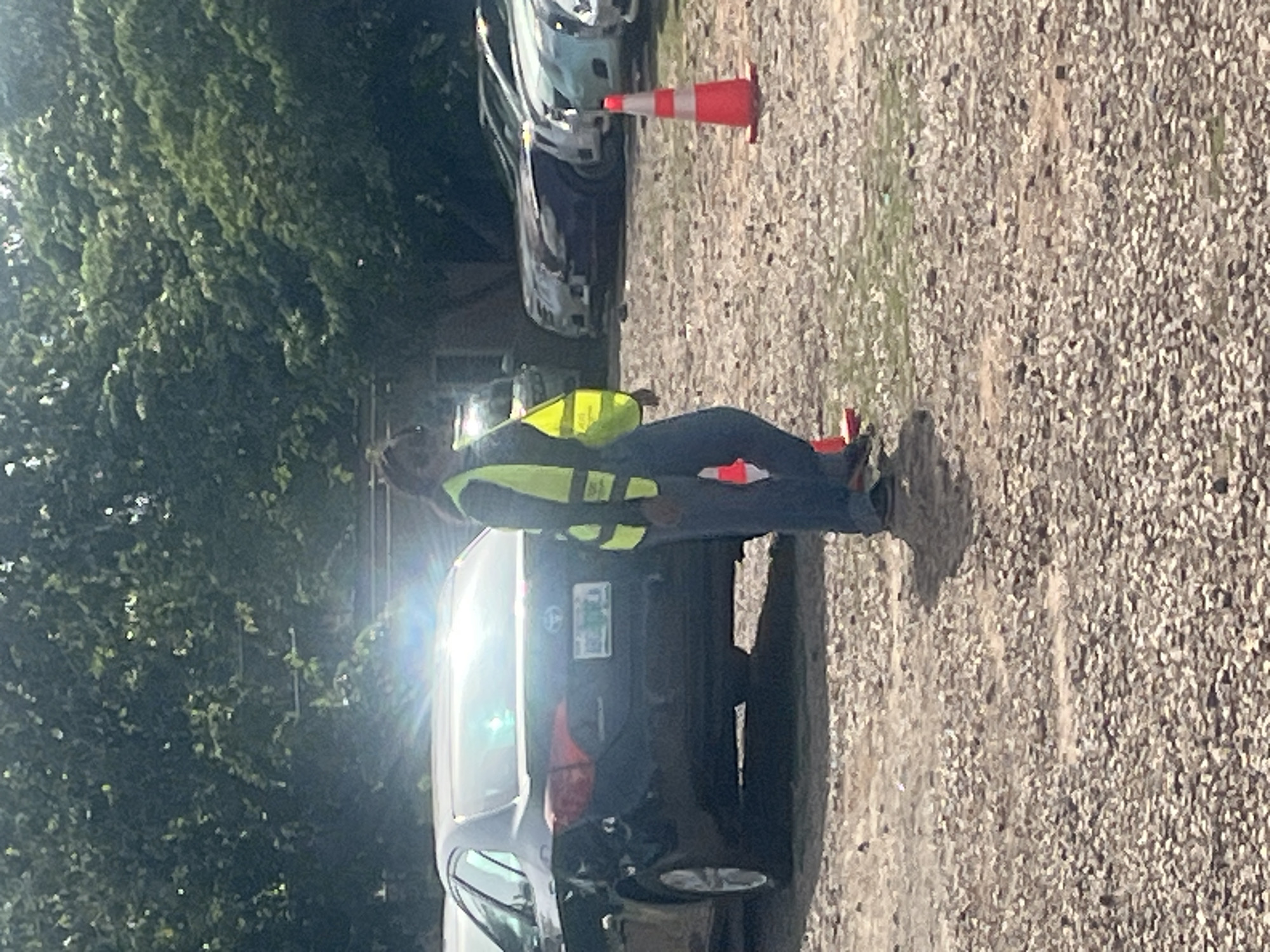 The image shows a man wearing a yellow reflective vest standing beside a dark-colored car in what appears to be a parking lot or gravel area. An orange traffic cone is near the car. There are trees in the background. The location is Galadimawa Village, Nigeria.