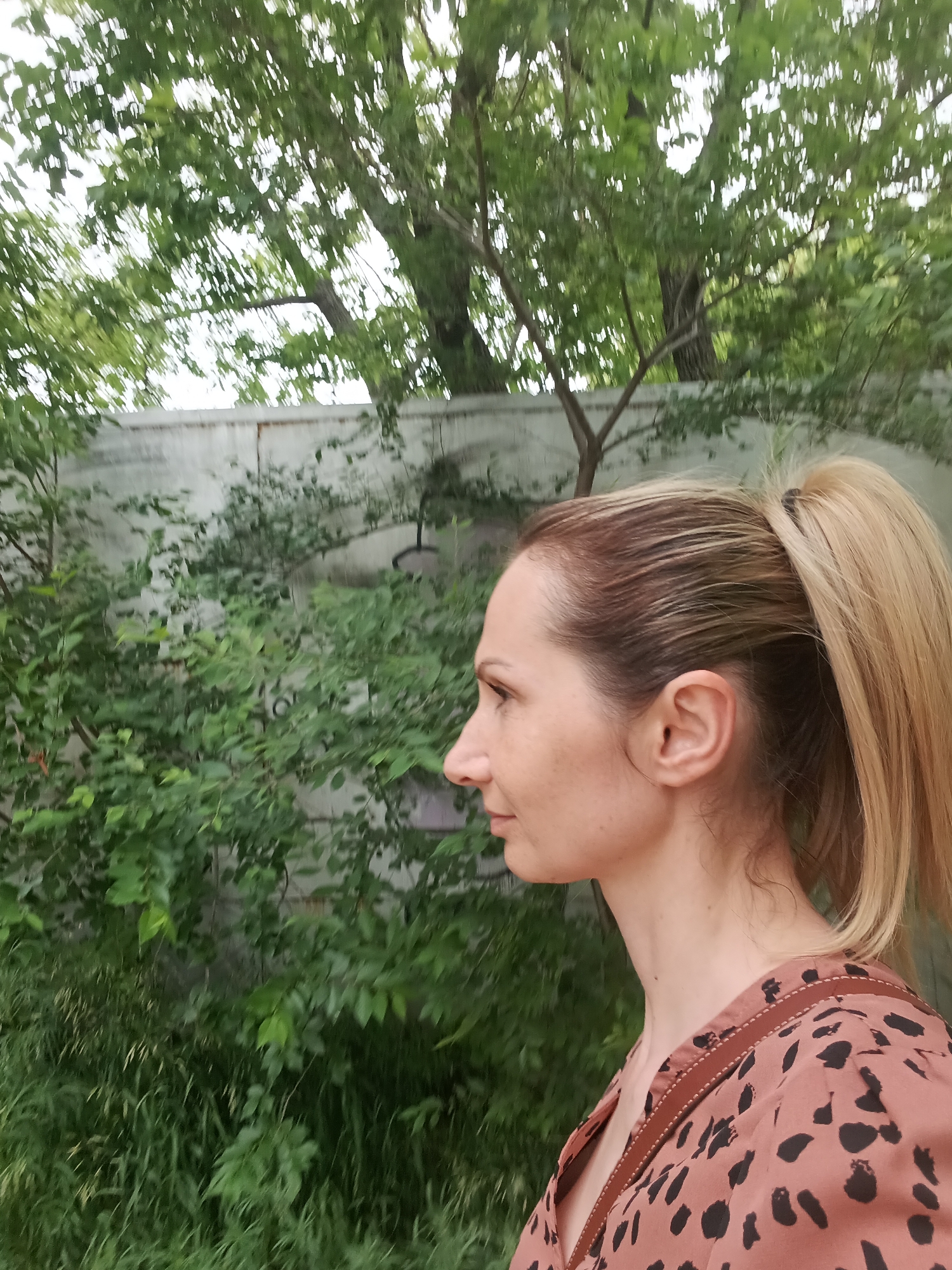 The image depicts a woman in profile, facing left, against a backdrop of greenery and a concrete wall. Her blonde hair is pulled back into a ponytail. She is wearing a rust-colored shirt with a black spotted pattern and brown trim. The environment appears to be outdoors, with trees and bushes visible. The concrete wall suggests an urban or semi-urban setting. The lighting indicates it is daytime. The location is Komsomolsk-on-Amur, Russia.