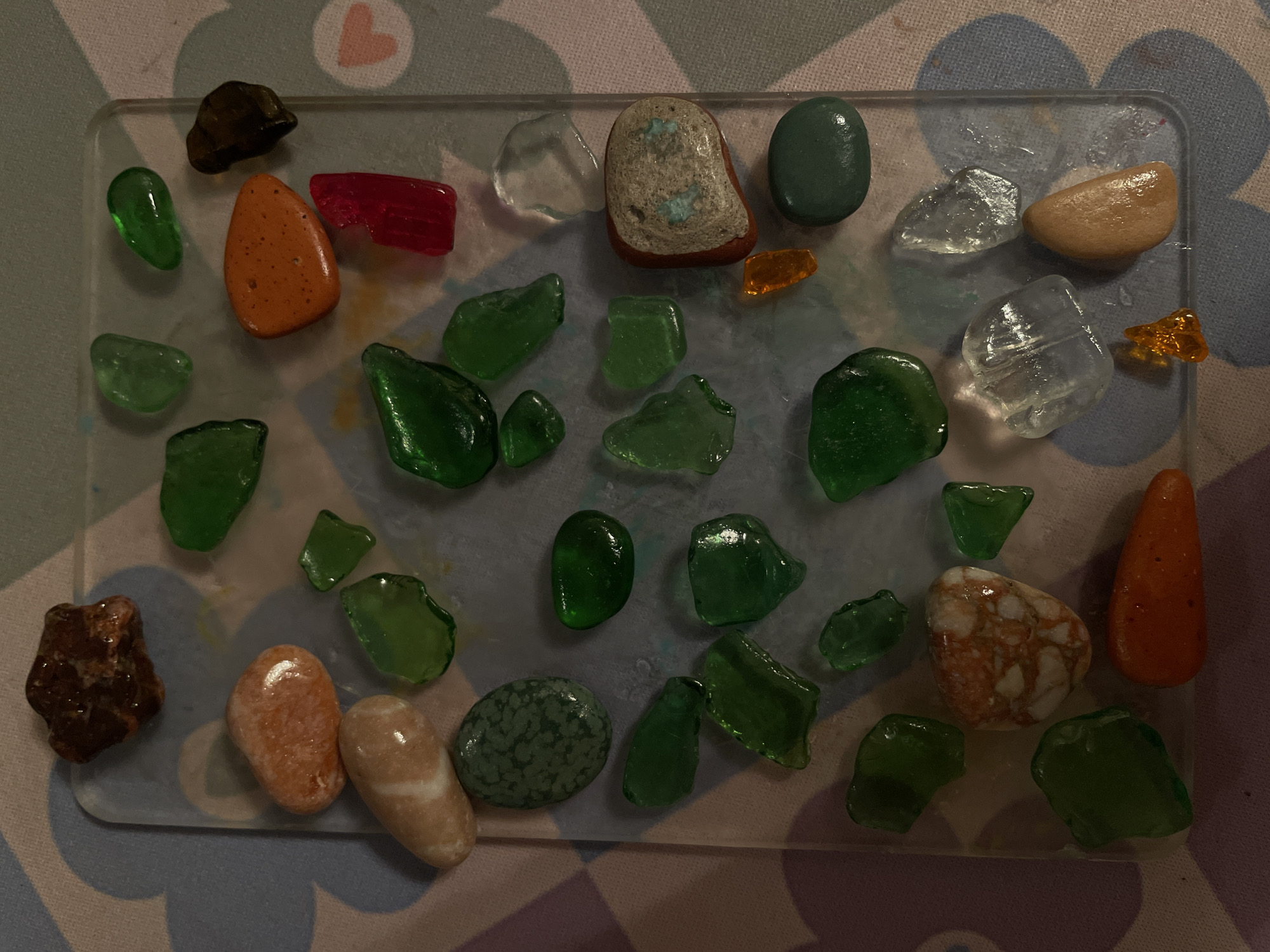The image shows a collection of smoothed glass and rocks, arranged on a clear plastic tray. The collection includes green glass shards of varying sizes and shapes, along with various stones, one of which is speckled. There is also a piece of red glass and a few pieces of orange glass. The tray rests on a patterned surface. There is no indication of location, time, setting, person, or action. Vracar Urban Municipality cannot be determined from the image.