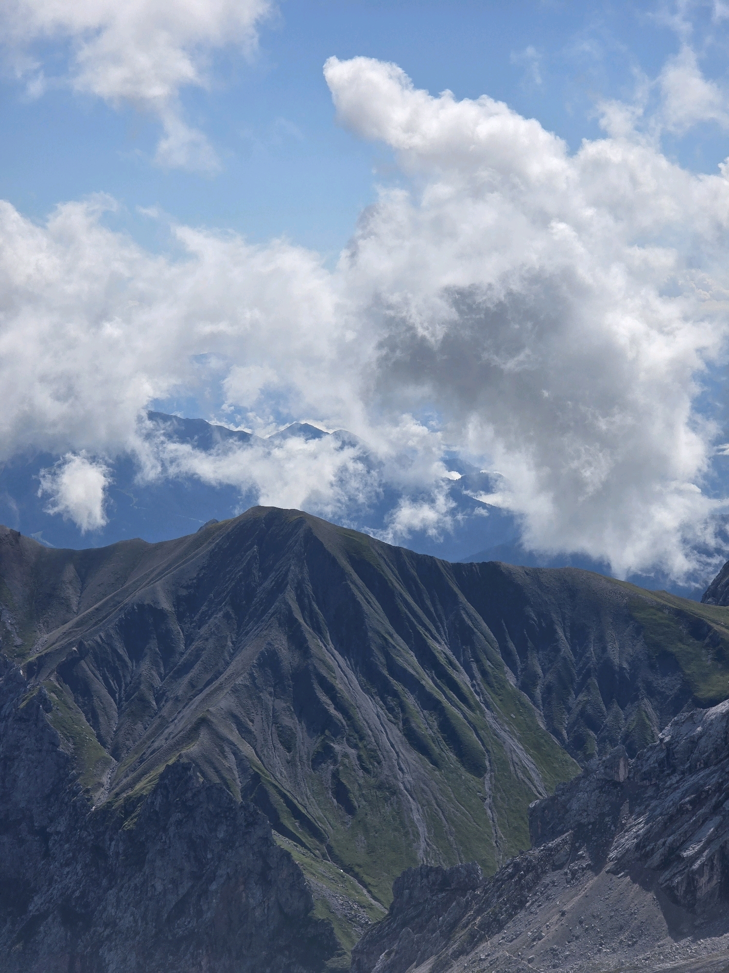 The image depicts a mountainous landscape with a dramatic sky. In the foreground, dark, rugged mountain slopes dominate the view. These slopes are characterized by steep inclines, deep ravines, and patches of green vegetation clinging to the rocky surfaces. The texture of the rock appears rough and weathered.
In the midground, additional mountain ranges are visible, appearing softer and more distant, with some peaks possibly snow-capped or covered in lighter-colored rock. These ranges are partially obscured by a layer of white, fluffy clouds.
The sky above is a vibrant blue, dotted with numerous clouds of varying sizes and densities. Some clouds are brightly lit, suggesting sunlight, while others have darker undersides, indicating depth and shadow. The clouds create a dynamic and somewhat dramatic atmosphere over the mountains.
The scene is entirely natural, with no human presence or man-made objects visible. The environment is a high-altitude mountain region, characterized by its rugged terrain and expansive sky. The lighting suggests it is daytime, likely mid-morning or afternoon, given the brightness of the clouds and the clear sky.