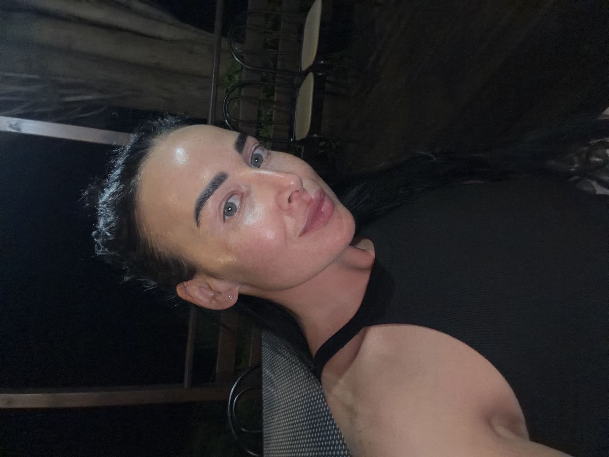 In a nighttime outdoor setting, a woman with dark hair pulled back from her face is captured in a close-up selfie. She wears a black, ribbed tank top and has light-colored eyes and fair skin with freckles. Her head is tilted slightly to her left, and she is looking directly at the camera with a faint smile. The background is dark, with some out-of-focus chairs visible, suggesting she is seated at a table in a restaurant or patio area. The lighting in the image appears to be artificial, casting a warm glow on her face. The location is identified as a restaurant terrace in Sochi, Russia.