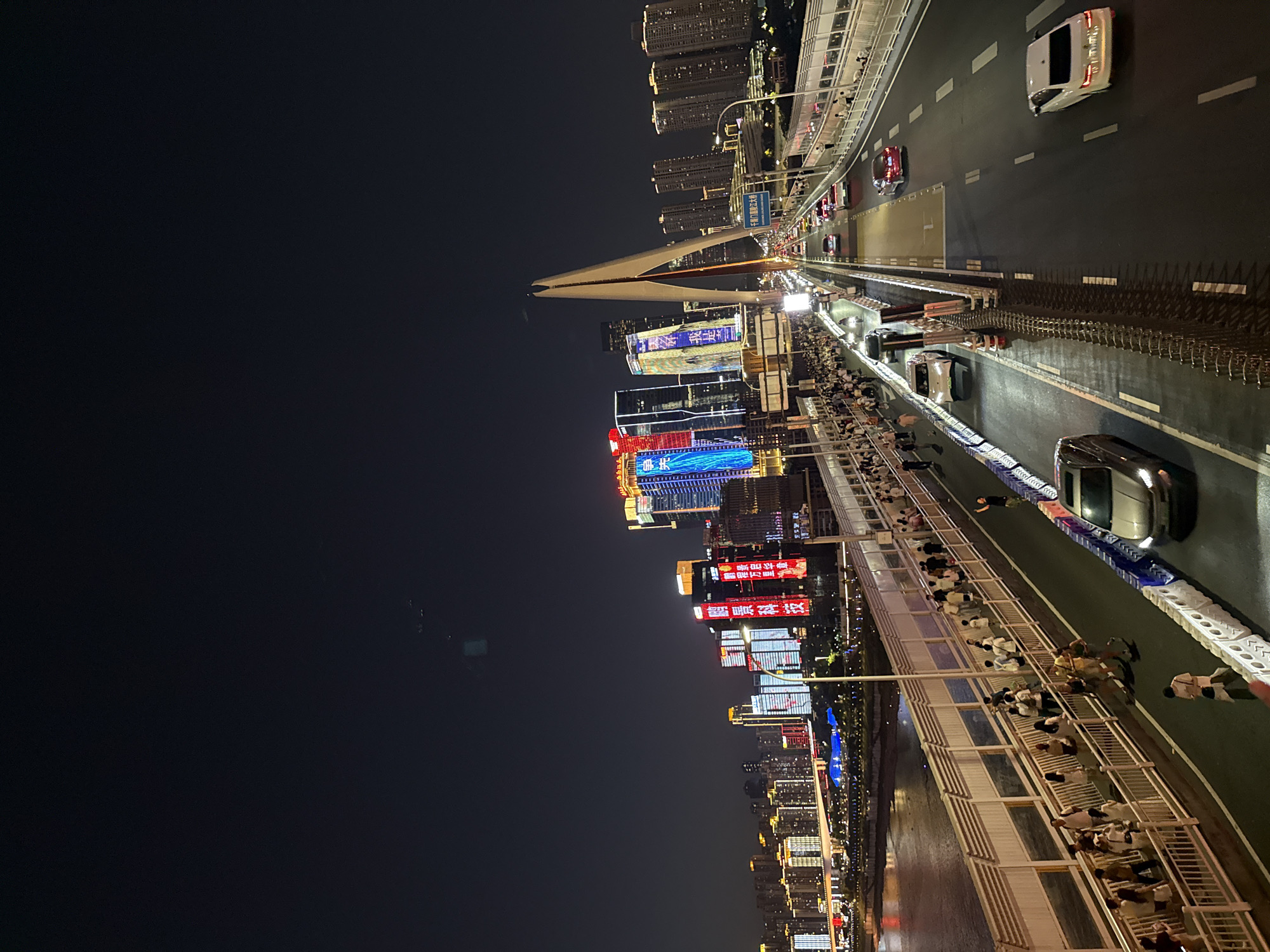 An elevated, long-shot, night-time photograph captures the cityscape of Yuzhong District, China. The foreground features a busy bridge with a pedestrian walkway on the left and multiple lanes of vehicle traffic on the right. Numerous pedestrians are walking on the designated walkway, while several cars are driving on the bridge. The bridge spans across a body of water, visible on the far left. In the background, a densely populated urban area is illuminated by the colorful lights of skyscrapers and buildings, forming a vibrant skyline. The sky is dark and clear. A prominent modern bridge pylon with a unique, angular design is centrally located in the mid-ground, with the bridge extending into the distance. The overall scene depicts a lively urban environment at night.
