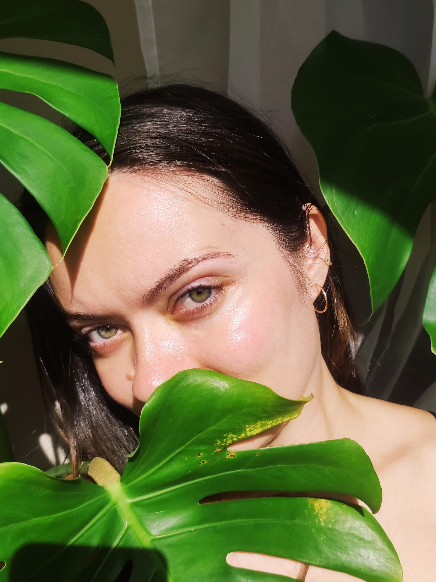 A young woman with dark hair and light skin is partially obscured by large, green Monstera plant leaves. Her face is in focus, with her green eyes looking directly at the viewer. Part of her nose and mouth are hidden behind a leaf. She is wearing small gold hoop earrings and a smaller earring higher up on her ear. Sunlight highlights her cheek. The background is blurred, showing a hint of a white curtain or wall. There is no indication of this photo being taken in Chaikovsky, Russia.