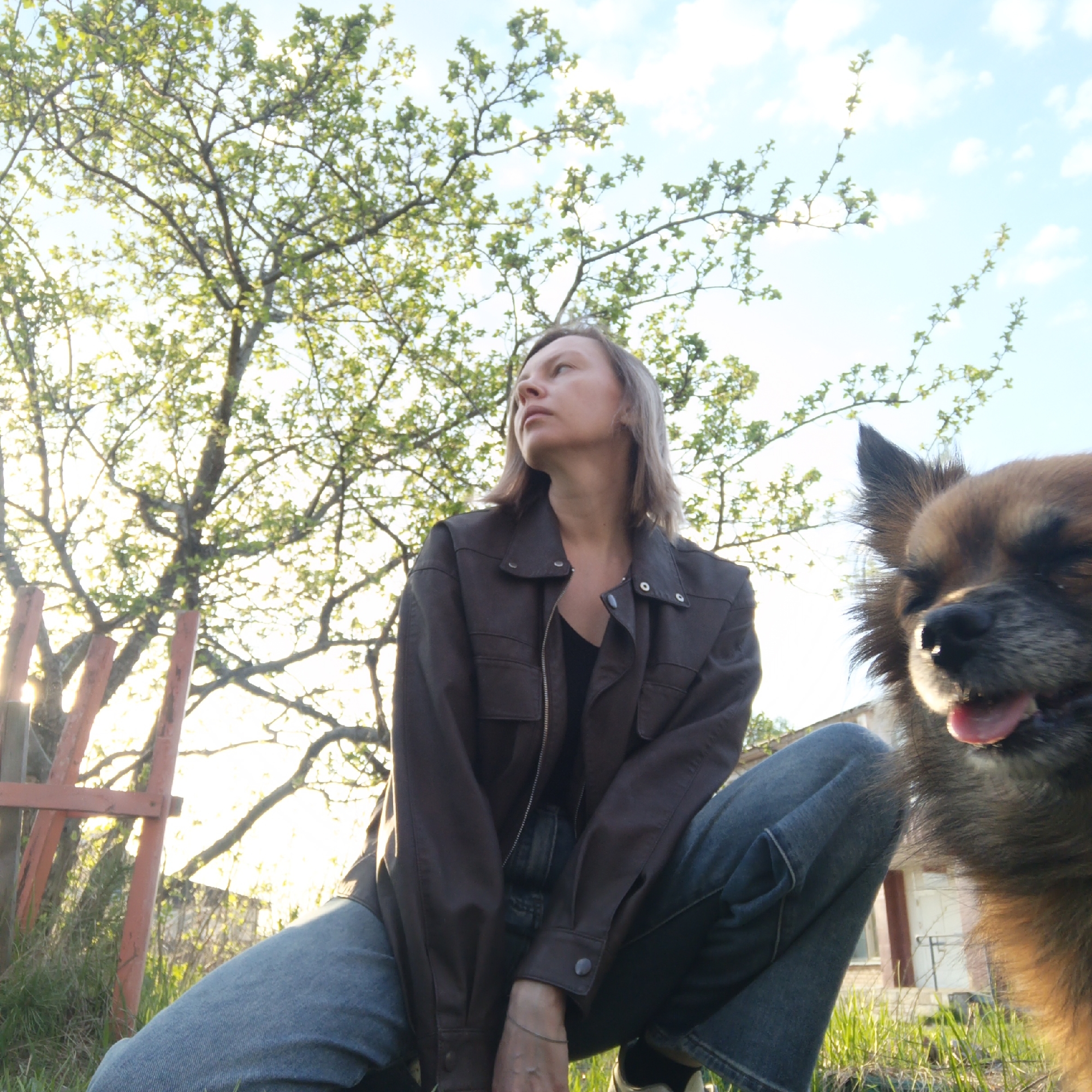 The image shows a woman crouching in an outdoor setting. She wears a brown jacket, a black top, and blue jeans, and has short blonde hair. She's looking to her left, away from the camera. To her right is a brown dog with its tongue hanging out. Behind her is a tree with young leaves against a blue sky with clouds. In the background, a building with a red fence is visible, and the overall scene suggests an outdoor, potentially residential area in Chaikovsky, Russia, given the prompt's specification. The presence of grass underfoot indicates a yard or garden setting.