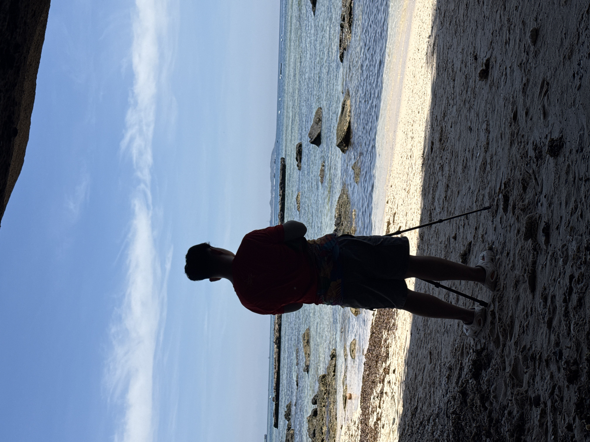 A person is standing on a sandy beach, facing the ocean, in Lý Sơn Special Administrative Region, Vietnam. The person is wearing a red t-shirt and dark shorts. They are standing behind a tripod, suggesting they may be taking photographs or filming. The beach is covered in sand and some scattered seaweed. The ocean is visible in the background with clear, calm water and several rocks emerging from the sea. The sky is blue with some wispy white clouds. The scene is partially shaded by an overhang, likely a rock formation.