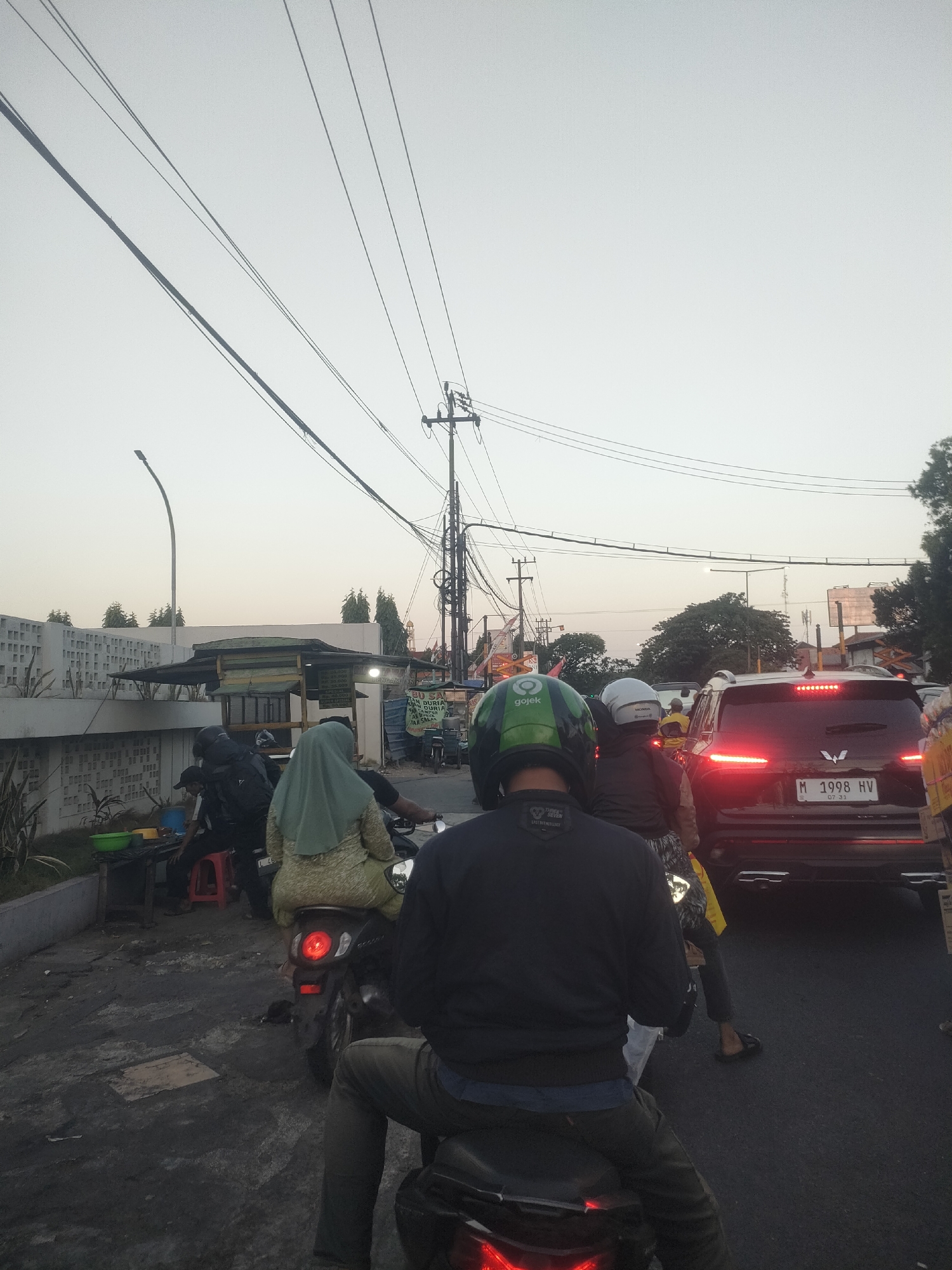 The image depicts a traffic scene in Surabaya, Indonesia, likely during the late afternoon or early evening due to the soft lighting and clear sky. Several motorcycles and a car are stopped, indicating a traffic jam. The foremost subject is a person on a motorcycle wearing a dark blue sweater and grey pants, facing away from the camera. To their left, another motorcycle carries two individuals; the passenger is wearing a light green hijab and patterned top. Further back, a black SUV with illuminated taillights is visible, displaying a license plate that reads "M 1998 HV 07 31". The surrounding environment includes utility poles with numerous electrical wires crisscrossing overhead, a lamppost, a low-rise building with a patterned facade, and a small roadside stall with a dark green awning. Vegetation, including trees, can be seen in the background. The ground is paved, appearing somewhat wet or dirty.