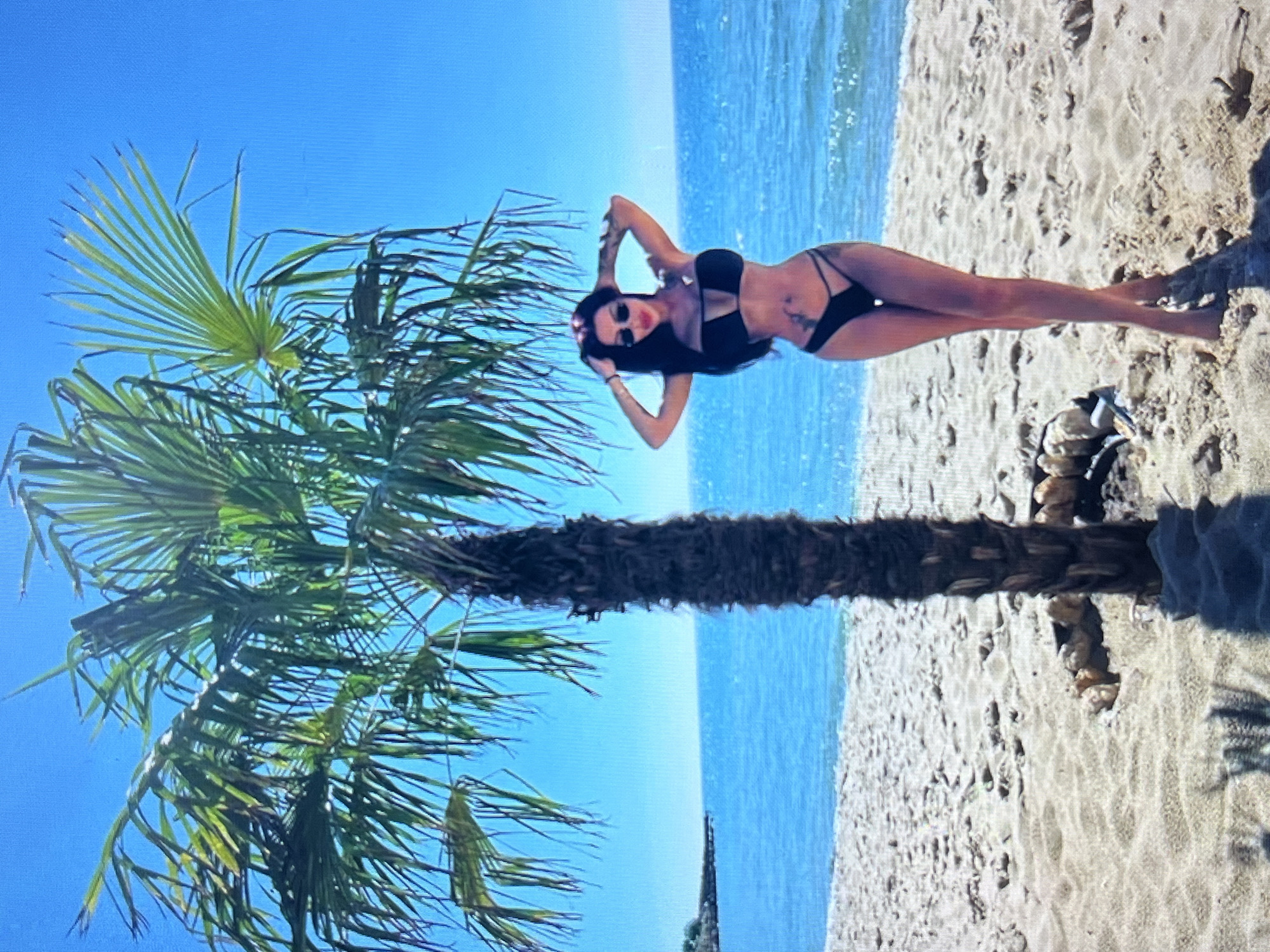 A full-body, eye-level, candid shot captures a woman standing on a sandy beach. She is wearing a black bikini and sunglasses, with her hands placed on her head. Behind her, a palm tree stands tall, and the ocean stretches out to the horizon under a clear blue sky. The woman appears to be posing for the camera. No specific landmarks or elements indicate the location is in Vratsa, Bulgaria.