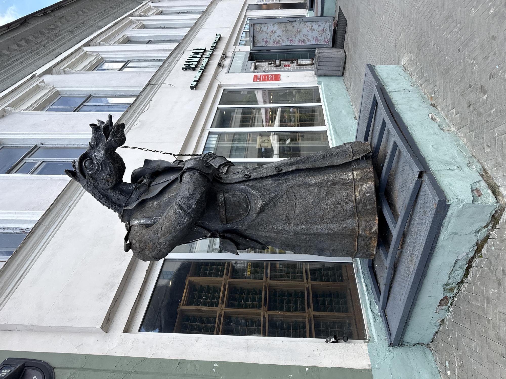 The image captures a street scene in Kazan, Russia, featuring a sculpture of a horse-headed figure dressed in a trench coat. The sculpture is positioned in front of a building with large windows and an entrance. One window displays stacked wooden crates filled with bottles, suggesting a shop or storage area. Above the entrance, a sign reads "САМОГОН" and "АНТИЛКОГОЛЬНЫЙ МУЗЕЙ С ДЕГУСТАЦИЕЙ," indicating an "Antialcohol Museum with Tasting." A red sign on the door displays operating hours. The sculpture is placed on a metal platform, which is situated on a light blue, weathered concrete base. The surrounding pavement is made of grey brick laid in a herringbone pattern. The building's facade is white with architectural detailing around the windows. A trash receptacle is located near the entrance. The sky is visible in the background, suggesting an outdoor setting during daylight.