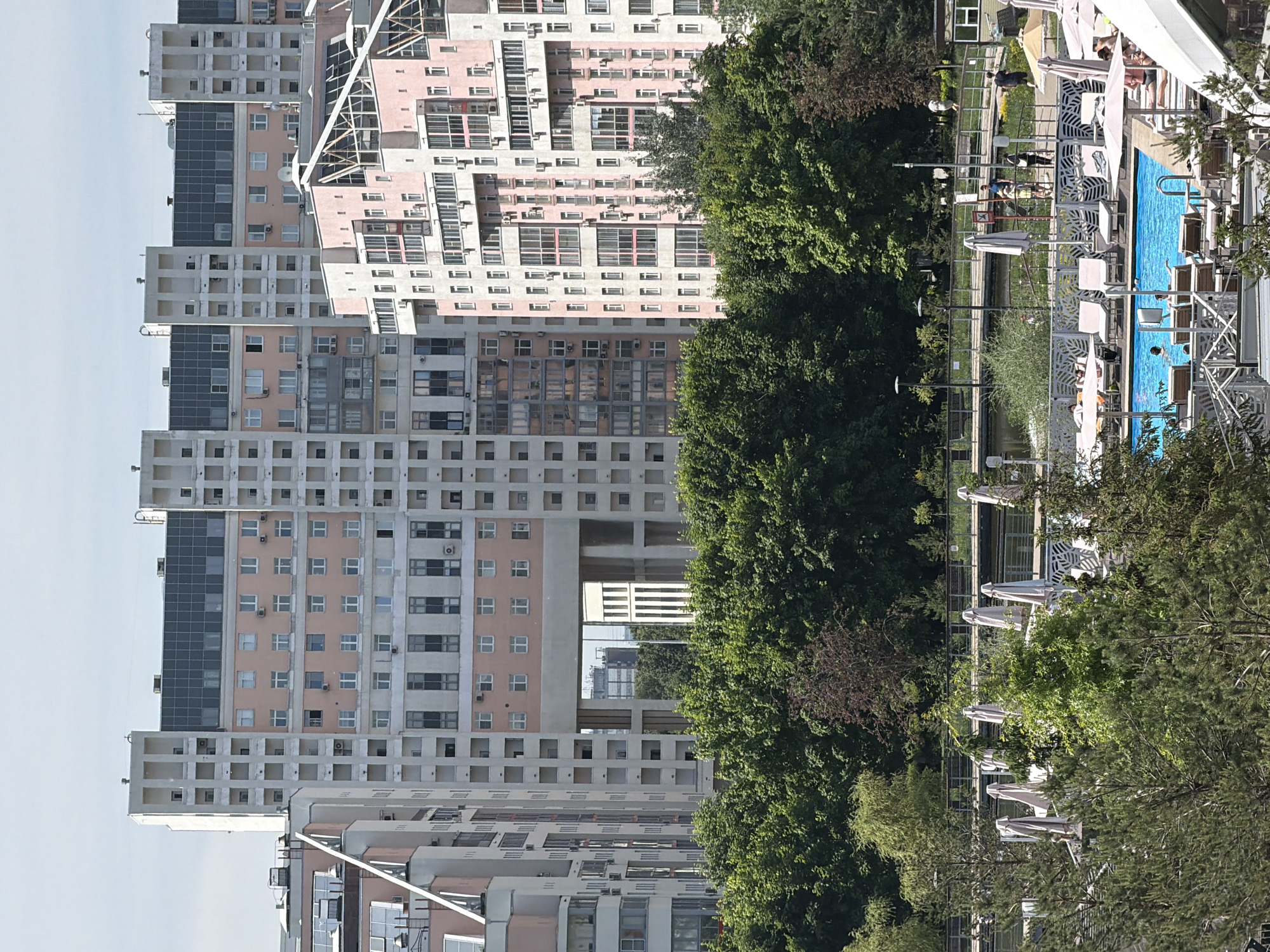 This detailed image depicts a modern residential and recreational scene in Khimki, Russia, under a clear, light blue sky.
In the background, a complex of multi-story residential buildings dominates the skyline. These structures primarily feature facades of light grey/white and a distinct salmon-pink color. The central and left-most large buildings are predominantly light grey with numerous square and rectangular windows, many displaying individual air conditioning units. The central building exhibits a unique architectural feature: a large, open rectangular cut-out at mid-height, through which distant structures and greenery are visible. The roofs of several buildings are dark, possibly covered with solar panels or a dark roofing material. To the right, another large residential building is prominently salmon-pink, characterized by numerous small, uniform windows and several enclosed or open balconies, many also fitted with air conditioning units. Adjacent structures also show a mix of light grey and pink elements, with varying window and balcony designs.
A dense band of lush green deciduous trees forms the middle ground, effectively separating the urban residential blocks from the foreground leisure area.
The foreground reveals a recreational complex centered around a bright blue swimming pool. The pool is rectangular, and several individuals are visible swimming or wading in its waters. Surrounding the pool is a light-colored paved deck furnished with numerous white sun loungers, some of which are occupied by people relaxing. Multiple closed, light-colored beach umbrellas stand upright around the pool area, with a few open ones also visible. White, modern-styled outdoor furniture and structures, possibly part of a cafe or club facility, are partially visible in the bottom right corner.
Behind the pool area and partially obscured by trees, a curved pedestrian bridge or elevated walkway spans a narrow body of water, possibly a canal or pond. Several people are walking along this bridge and the paths surrounding it. Streetlights, metal railings, and park benches are also discernible along the walkway. The overall scene suggests a sunny day with active leisure pursuits.