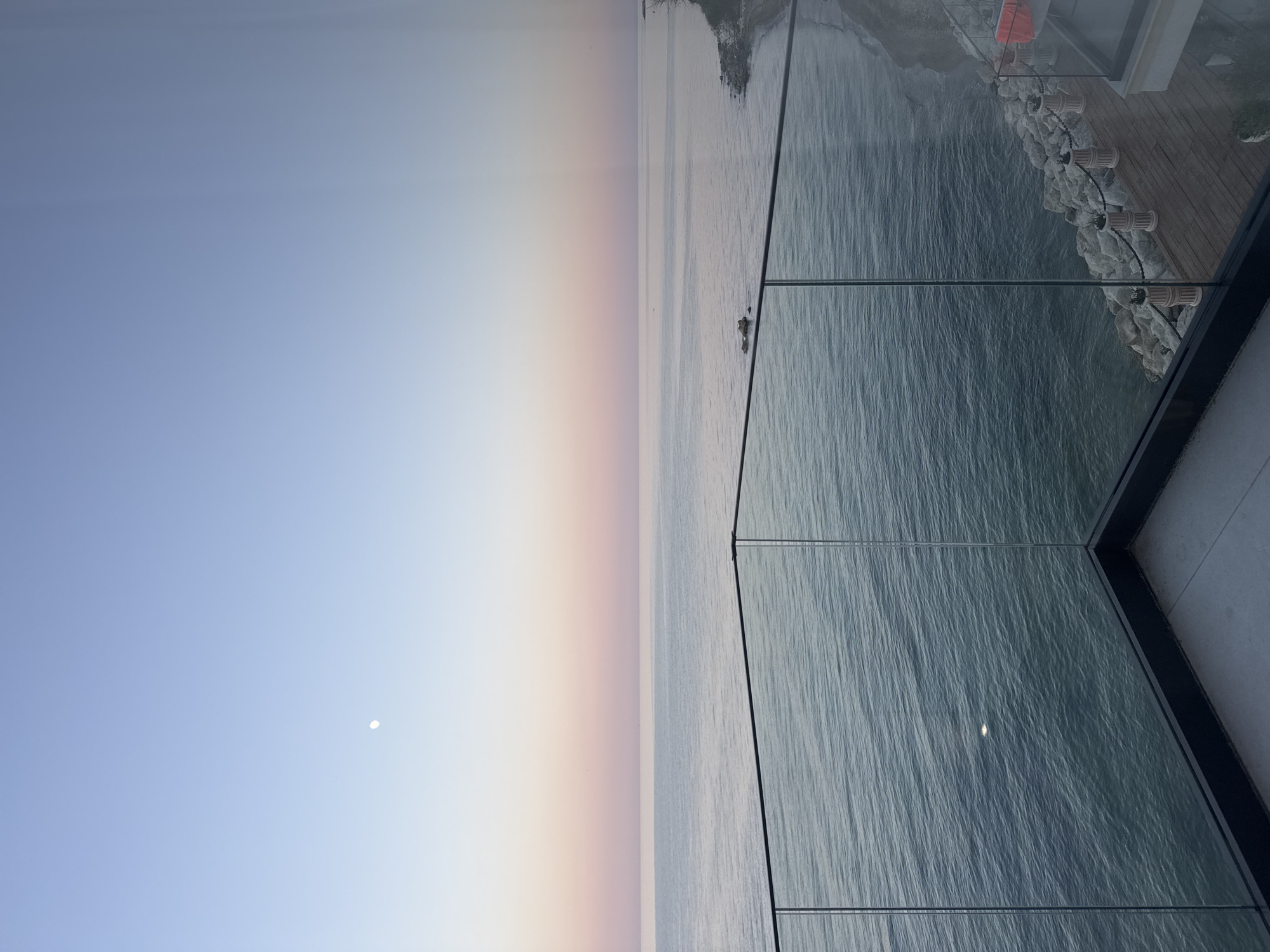 This image depicts a tranquil scene overlooking the sea, likely during twilight or dawn given the soft, gradient hues of the sky transitioning from a pale blue to a warm peach. The sea is calm, with gentle ripples reflecting the ambient light.
In the foreground, a modern architectural element, possibly a balcony or viewing platform, is visible. It features a glass railing that provides an unobstructed view of the seascape. The glass itself reflects the sky and the water, creating an ethereal effect. Beneath the glass, a wooden deck is laid out, bordered by a stone retaining wall. Several planters with decorative elements are arranged along the wall. A small boat is visible in the distance on the water.
The sky is mostly clear, with a small, bright circle that could be the moon or a distant light source visible in the upper left quadrant of the image. The overall mood is peaceful and serene, evoking a sense of quiet contemplation. The location is identified as Varna, Bulgaria. No text is visible in the image.