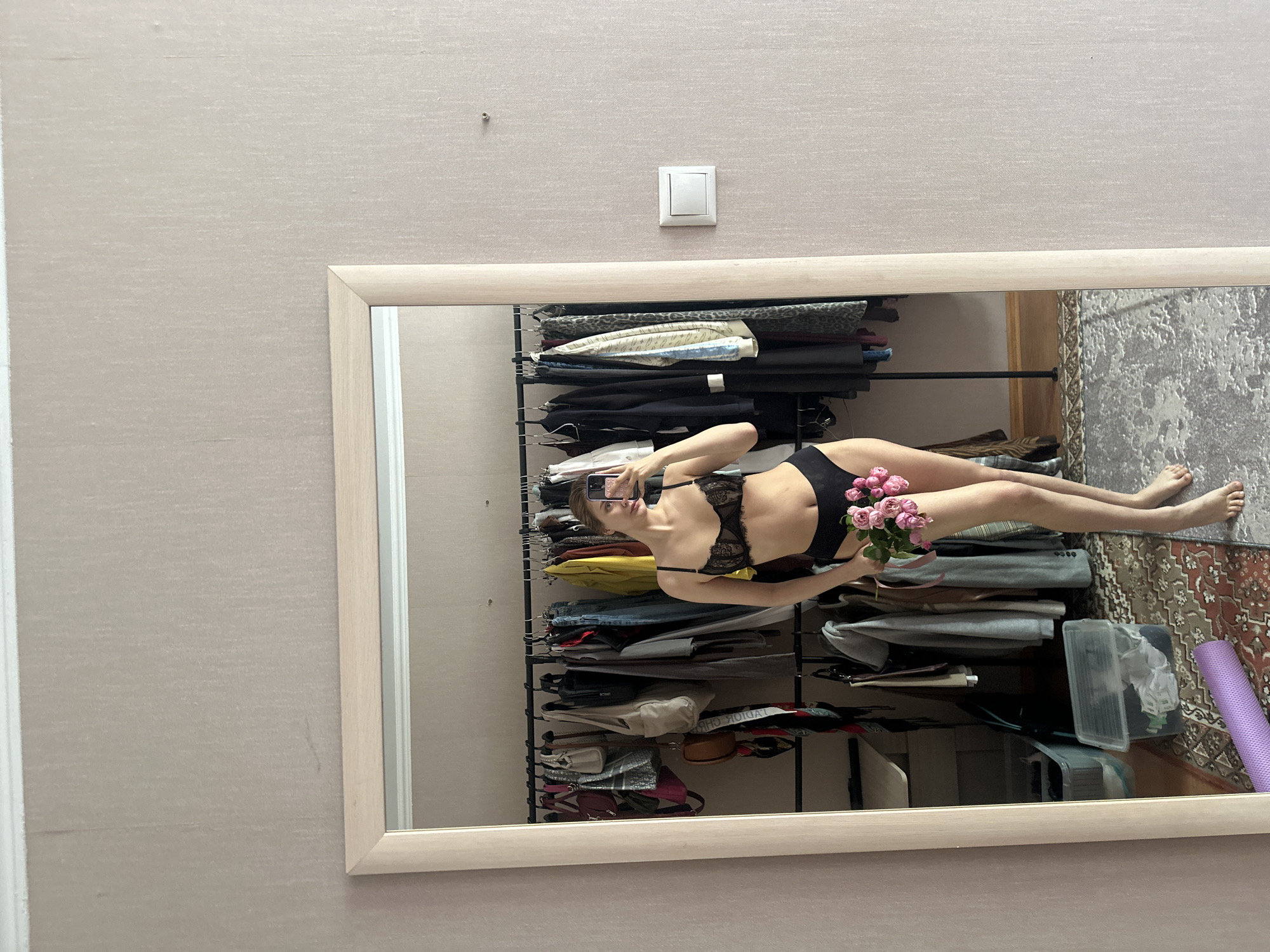 A woman is seen reflected in a mirror, taking a selfie with a smartphone. She is wearing black lingerie and holding a bouquet of pink roses. The reflection shows a bedroom environment with a clothes rack filled with various garments, a patterned rug on the floor, and a purple yoga mat. The room's decor includes a light switch on the wall, which is painted in a neutral tone. The location of the image is likely an apartment in Krasnoyarsk, Russia, given the style of the interior and the user's request.