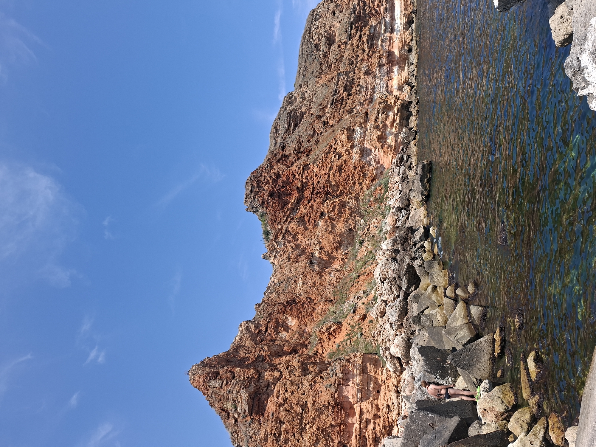 An image depicts a scenic coastal view with a person near the water and a prominent rock formation under a bright blue sky. The person, positioned on the left side of the frame, is a female wearing a dark bikini and light-colored footwear, standing on a rocky outcrop. She is looking towards the water. The environment features a rocky shoreline with various sizes of stones and boulders leading to clear, blue water. In the background, a large, rugged cliff face dominates the scene, characterized by its reddish-brown hue and layered geological structure. The sky is clear with scattered white clouds, indicating a sunny day. The location is identified as Balgarevo, Bulgaria.