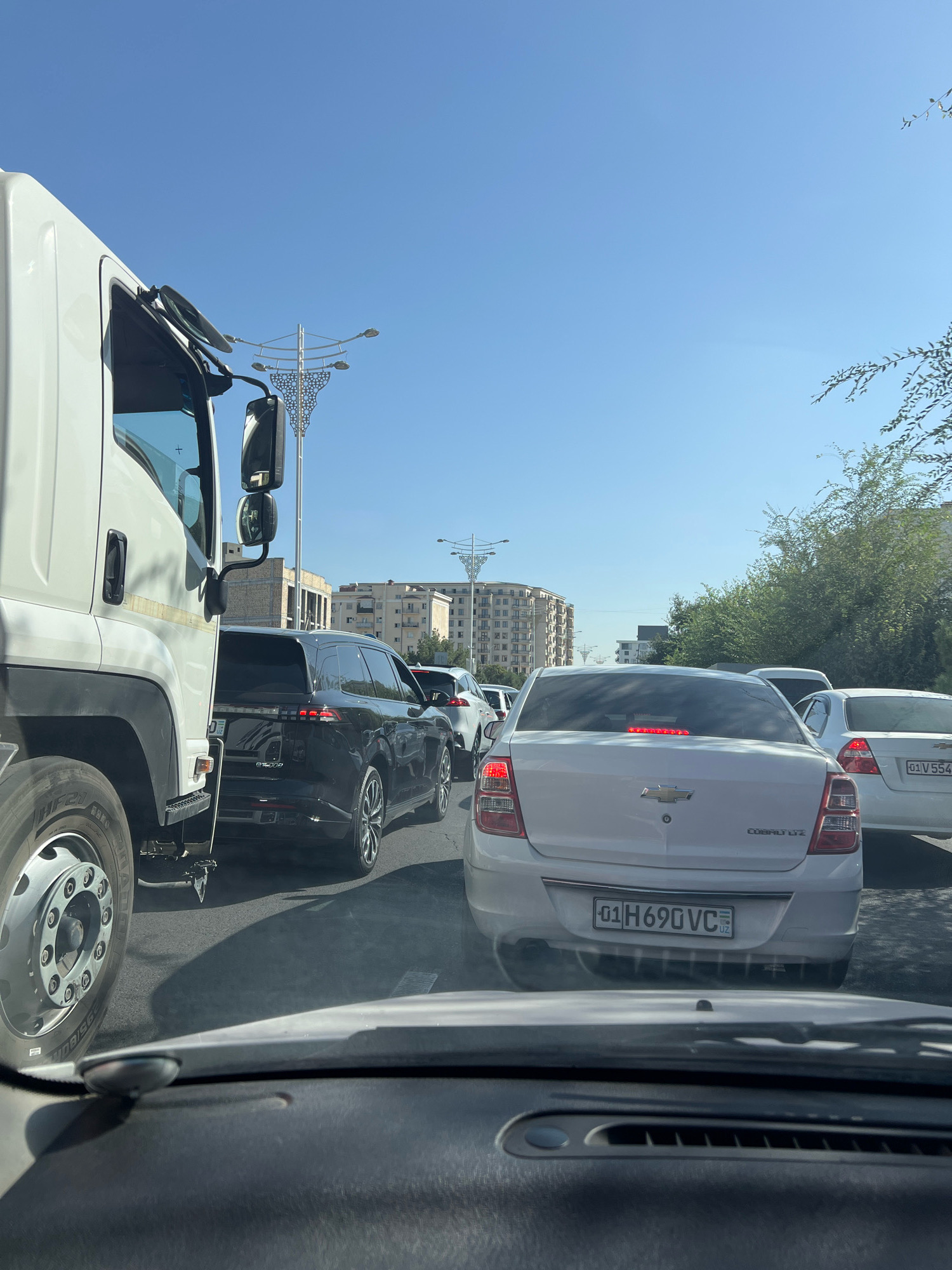The image depicts a daytime scene of heavy traffic congestion on a sunny day in Tashkent, Uzbekistan. The viewpoint is from inside a vehicle, looking forward. In the foreground, the dashboard of the vehicle is visible. Immediately ahead is the rear of a white Chevrolet Cobalt LYZ, displaying a license plate that reads "01 H690 VC" with a small Uzbek flag emblem. Behind this Chevrolet, another white car is visible, with a license plate reading "01 V 554". To the left of the Chevrolet is a black SUV, and further left, the front of a large white truck occupies a significant portion of the frame, with its wheel clearly visible. The background shows a street lined with trees and several multi-story buildings, characteristic of urban Tashkent. The sky is clear and blue. Streetlights with decorative elements are visible in the distance. The overall impression is a typical urban traffic jam during daylight hours.
