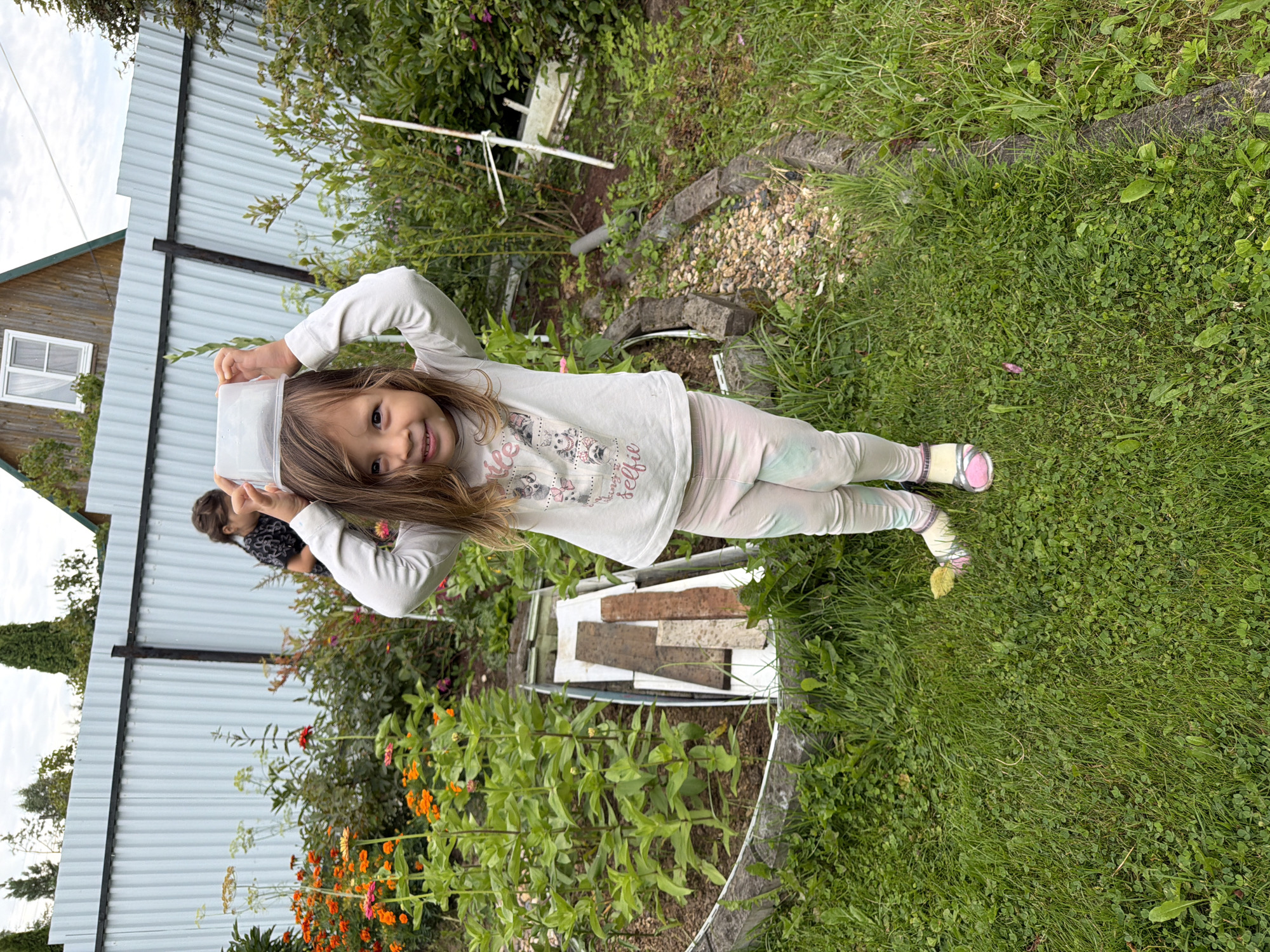 A young girl stands in a garden, holding a transparent plastic container on her head. She is wearing a long-sleeved shirt with a graphic print and light-colored leggings. The garden features lush green grass, a flowerbed with orange and pink flowers, and a light blue corrugated metal fence in the background. Behind the fence, a wooden house with a window is partially visible. Another child, a girl with dark hair pulled back, stands near the fence in the background. The scene appears to be in a rural or suburban setting, possibly a dacha or backyard.