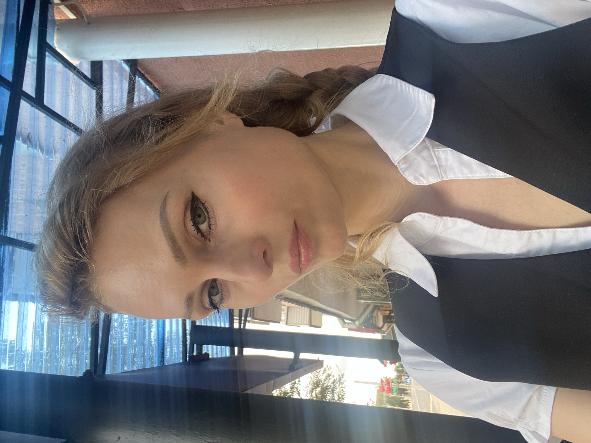 A fair-skinned woman with blonde hair in a braid, wearing a white collared shirt and a black vest, is photographed outdoors in Lida, Belarus. She is looking directly at the camera with a neutral expression. The background shows a street scene with buildings and trees, suggesting an urban environment. The lighting indicates it is daytime.