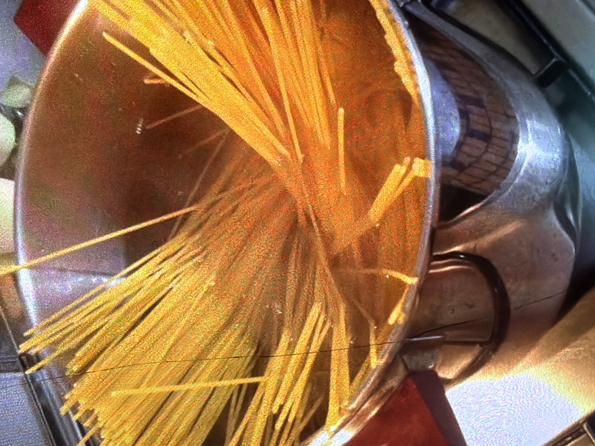 A close-up shot shows spaghetti in a pot of boiling water. The spaghetti is bright yellow and submerged in the water, with some strands extending above the surface. The pot appears to be made of stainless steel and is on a stovetop. The water is clear and there are some small bubbles indicating it is boiling. The lighting suggests an indoor environment, possibly a kitchen. The image does not provide any information about the city of Bukuru, Nigeria.