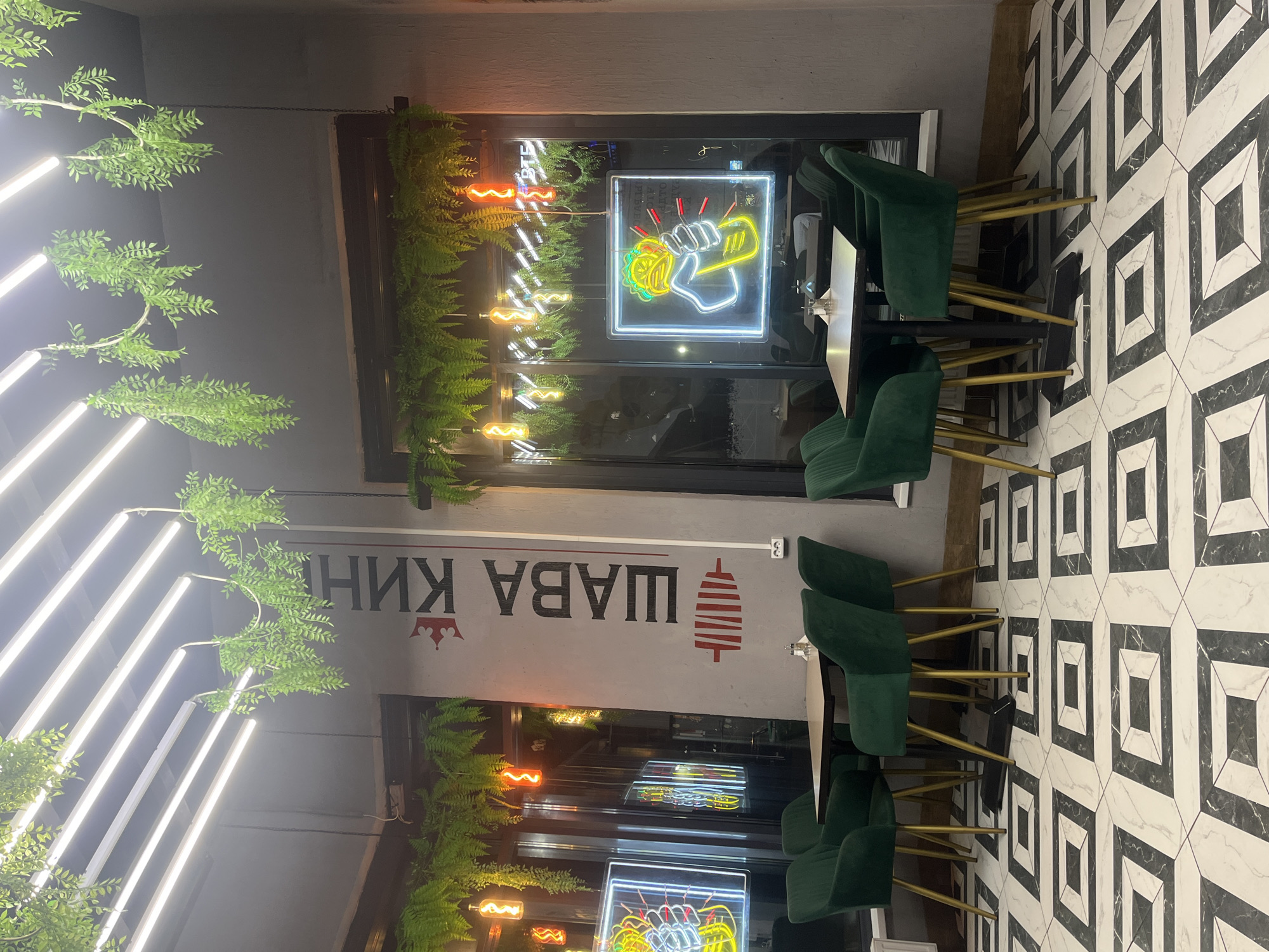 The image captures an indoor scene in what appears to be a restaurant. The flooring consists of black and white patterned tiles. Dark green upholstered chairs are arranged around tables. A window is adorned with green plants, with neon signs visible in the window's reflection. The text "SHAWARMA KING" is displayed prominently on the wall, in what appears to be the Russian language, along with a graphic image of shawarma. The scene has artificial lights that reflect in the window. Based on the text "SHAWARMA KING," the image suggests a fast-food establishment, possibly located in Saint Petersburg, Russia.