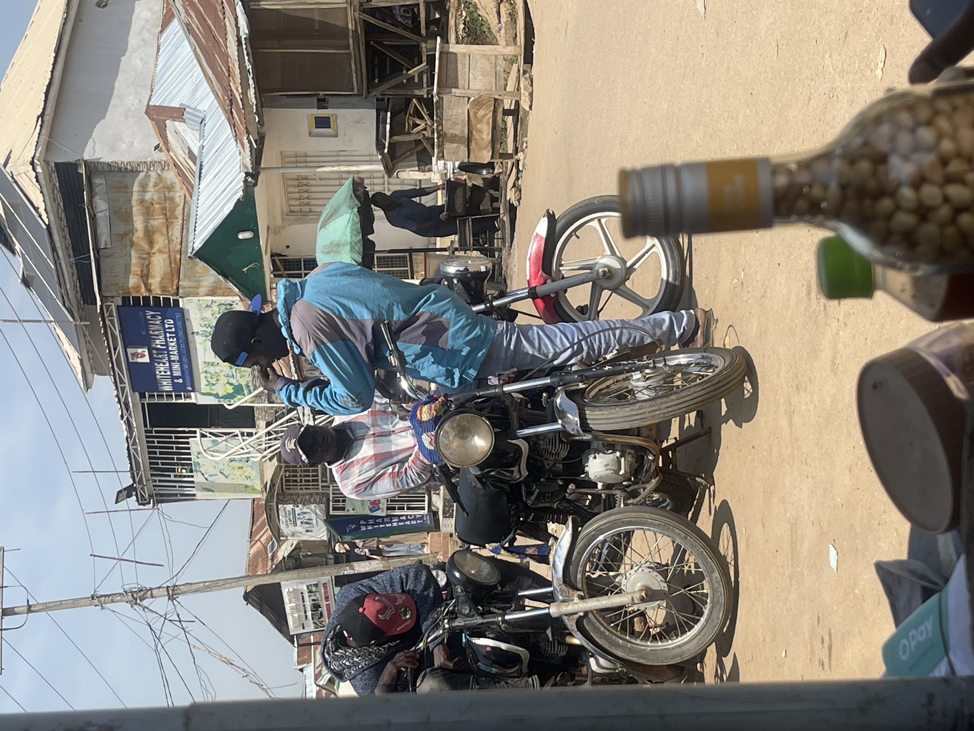 In an outdoor scene in Abuja, Nigeria, two men are standing next to motorcycles, with a third person seated on one of the motorcycles. One man in the foreground wears a blue and grey jacket and blue jeans. The second man, slightly behind and to the left, is dressed in a plaid shirt and a dark cap. The third individual, partially visible, is seated on a motorcycle and is wearing a dark hooded garment and a red cap. The motorcycles appear to be parked on a dirt road in front of a row of buildings. The buildings have corrugated metal roofs and some have signs, including one for "WHITEHEART PHARMACY & MINI-MARKET LTD". The overall environment suggests a commercial area, possibly a market or a street with small businesses. There are power lines visible overhead, and the sky is clear. In the foreground, a blurred bottle containing small, light-colored objects and another bottle with a green cap are partially visible, suggesting the image was taken from a close vantage point, possibly from behind a counter or through a window.