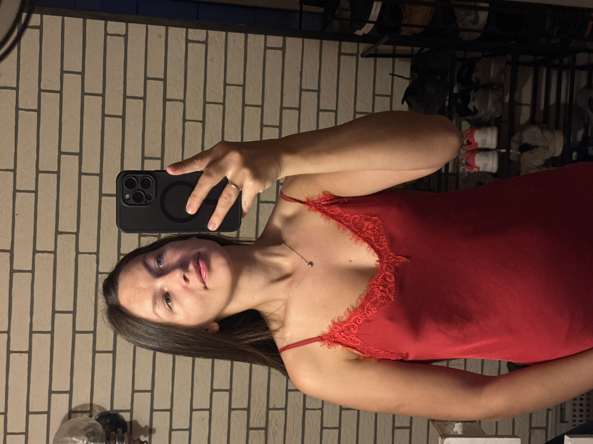 A young woman is depicted taking a mirror selfie indoors. She is positioned centrally in the frame, slightly angled, with her head tilted and her gaze directed towards her reflection. She has long, dark hair, light-colored eyes, and a calm, neutral expression.
She is wearing a red camisole or slip dress, detailed with intricate lace trim along the V-neckline. Around her neck, she sports a slender necklace with a small, round pendant, and a simple ring is visible on her left ring finger. In her right hand, she holds a dark-colored smartphone, featuring a distinct triple-camera module and a circular attachment on its case, extended as she captures the image.
The background shows a wall with a light-toned, brick-like pattern defined by dark grout lines. To the right, a multi-level shoe rack filled with various footwear, including sneakers, is partially visible. The lighting within the scene appears artificial and somewhat subdued, suggesting an interior setting, likely a private residence. No discernible text is present in the image.