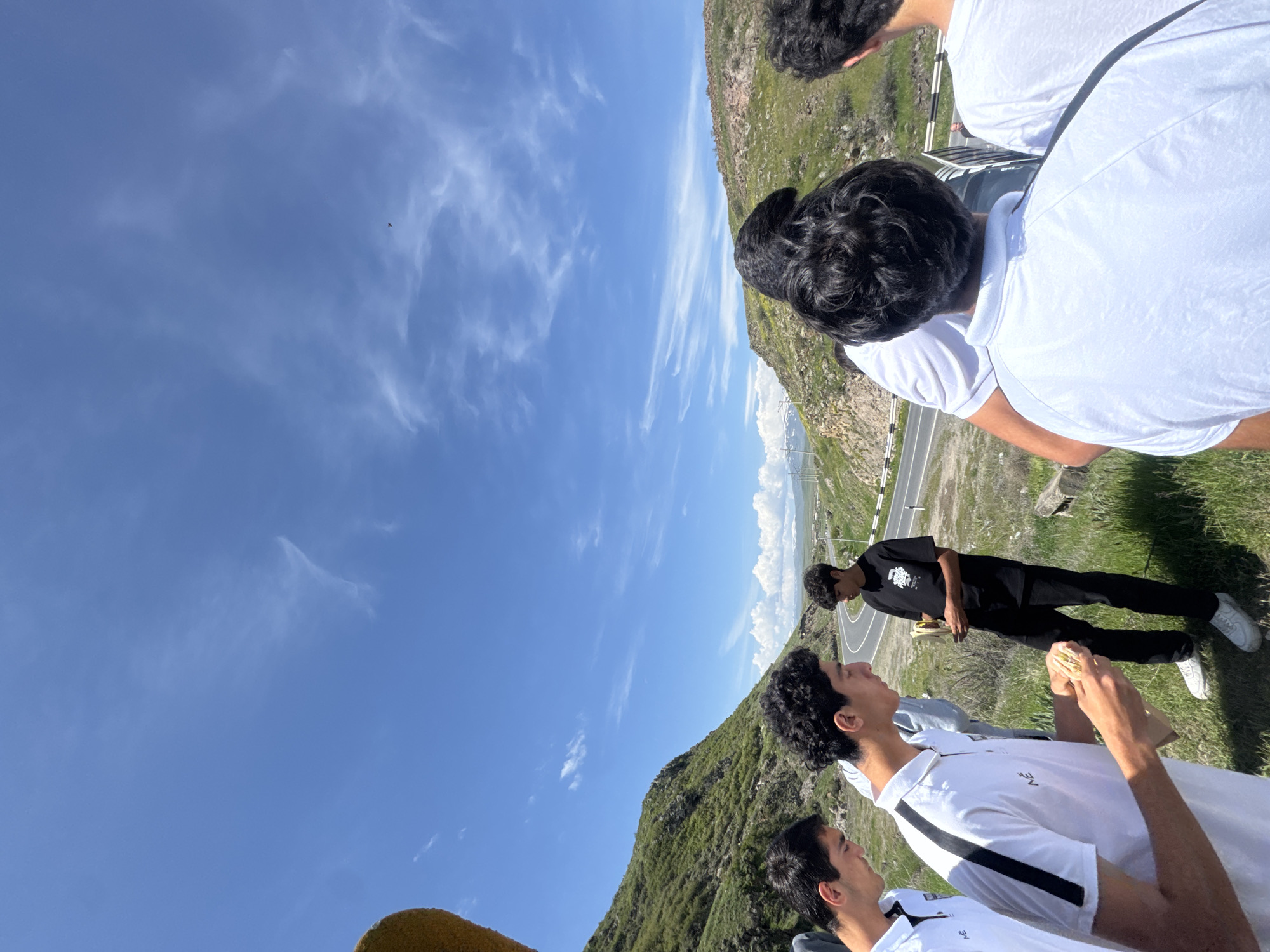The image captures a group of people in Bagravan, Armenia. Several people are present, some wearing white shirts and others in black. The backdrop features a mountain landscape and a clear blue sky with scattered clouds. A road winds through the mountain area. One person is holding food, seemingly a sandwich. The others appear to be looking at the landscape.