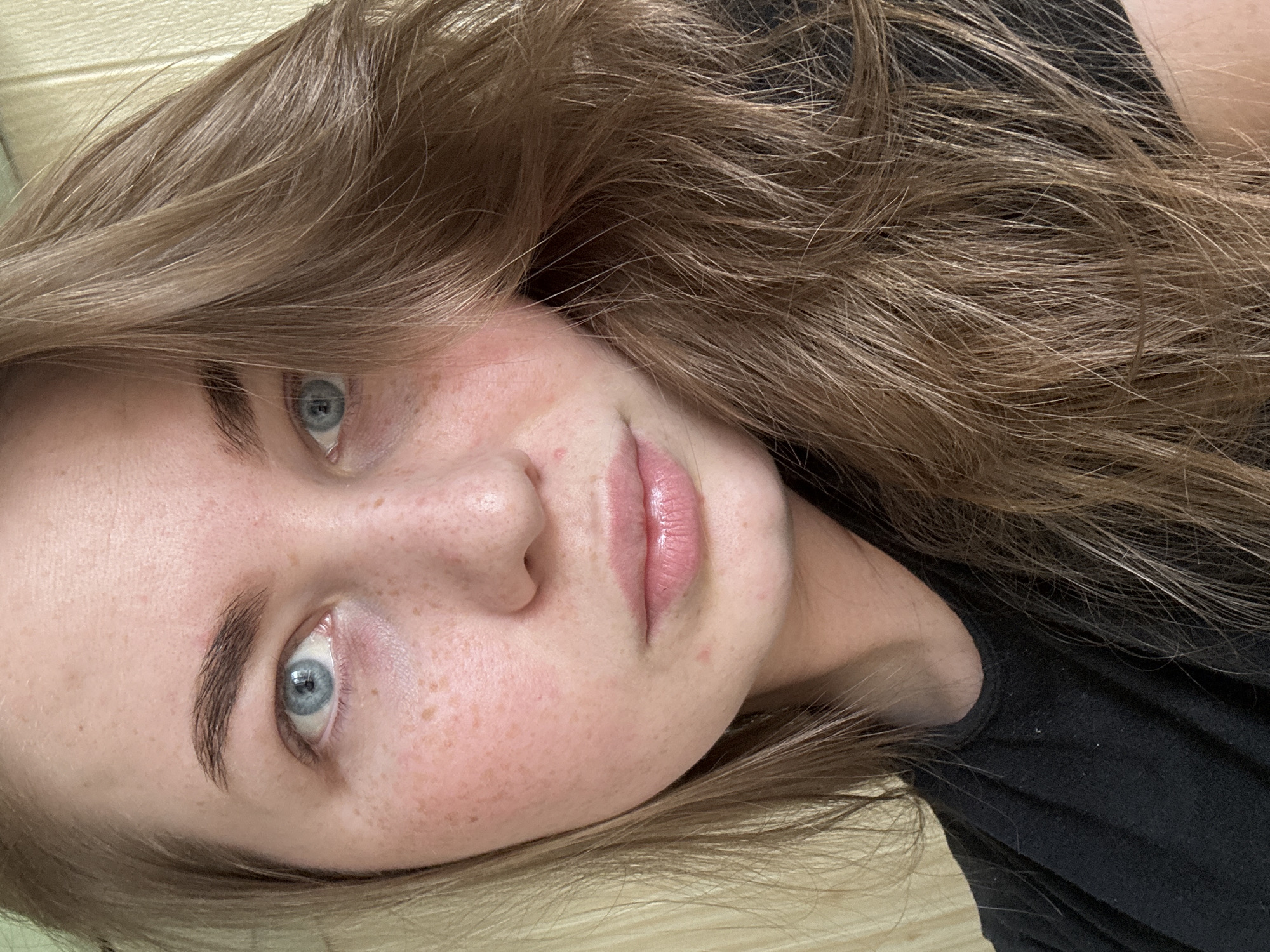 A close-up portrait features a person, likely a young female, looking directly at the camera with a neutral expression. The image is rotated 90 degrees clockwise, presenting the person's head tilted to the side.
The subject has light brown or auburn hair, appearing naturally wavy and falling around their face. Their skin is fair with numerous visible freckles across the cheeks and forehead. The eyes are a striking blue, with well-defined, natural-looking eyebrows. The lips are a soft pink, slightly parted, and appear moisturized or with a subtle gloss. A small portion of the nose is visible.
A black garment, likely a t-shirt or casual top, is visible around the neck and shoulder area on the right side of the frame. The background is a light-colored, textured surface, possibly wood paneling or a wall, suggesting an indoor setting. The lighting is soft and even, highlighting the facial features. No specific location cues are visible beyond the implied indoor environment, and there is no visible text. The overall mood is calm and direct.