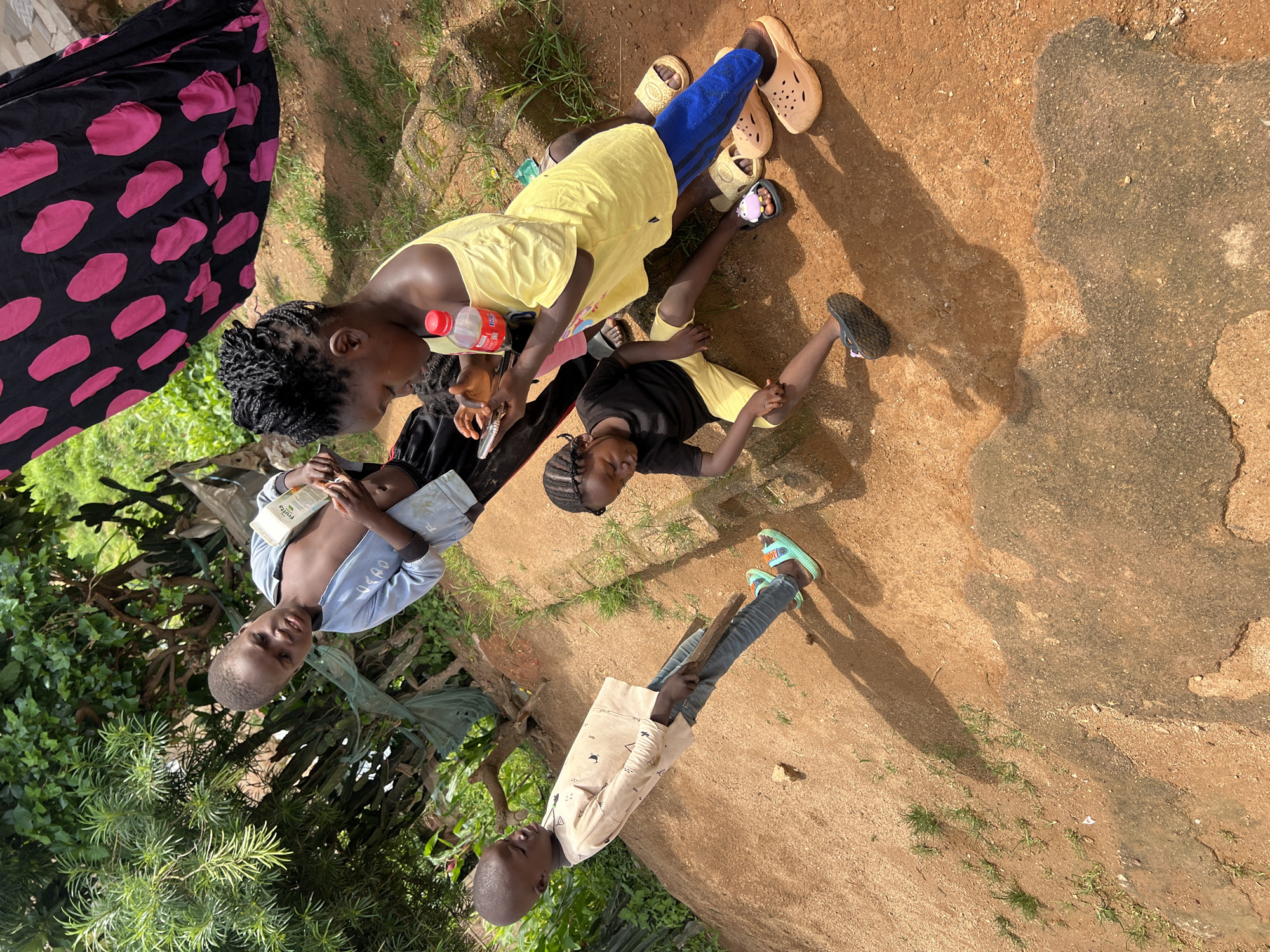 In the city of Jos, Nigeria, four children are gathered outdoors on a reddish-brown dirt ground. A large black and pink polka dot cloth is draped in the background.
On the left, a young boy wearing a light tan long-sleeved shirt and blue jeans stands leaning forward, holding a wooden stick. He wears mint green sandals.
In the center-left, a boy in a light blue zip-up jacket, open to reveal his bare chest, holds a white carton of juice.
To the right of the boy with the juice, a young girl with braided hair in a yellow off-the-shoulder top and blue pants sits on the ground, looking down at a smartphone she holds in her hands. A red bottle of Coca-Cola is beside her. She wears tan slip-on sandals.
In the front-center, a young child in a black t-shirt and yellow shorts sits with their legs outstretched. They are barefoot.
The environment consists of sparse grass and some greenery in the background, suggesting a rural or semi-rural setting. Shadows from the children and surrounding vegetation are cast on the ground, indicating a sunny day.