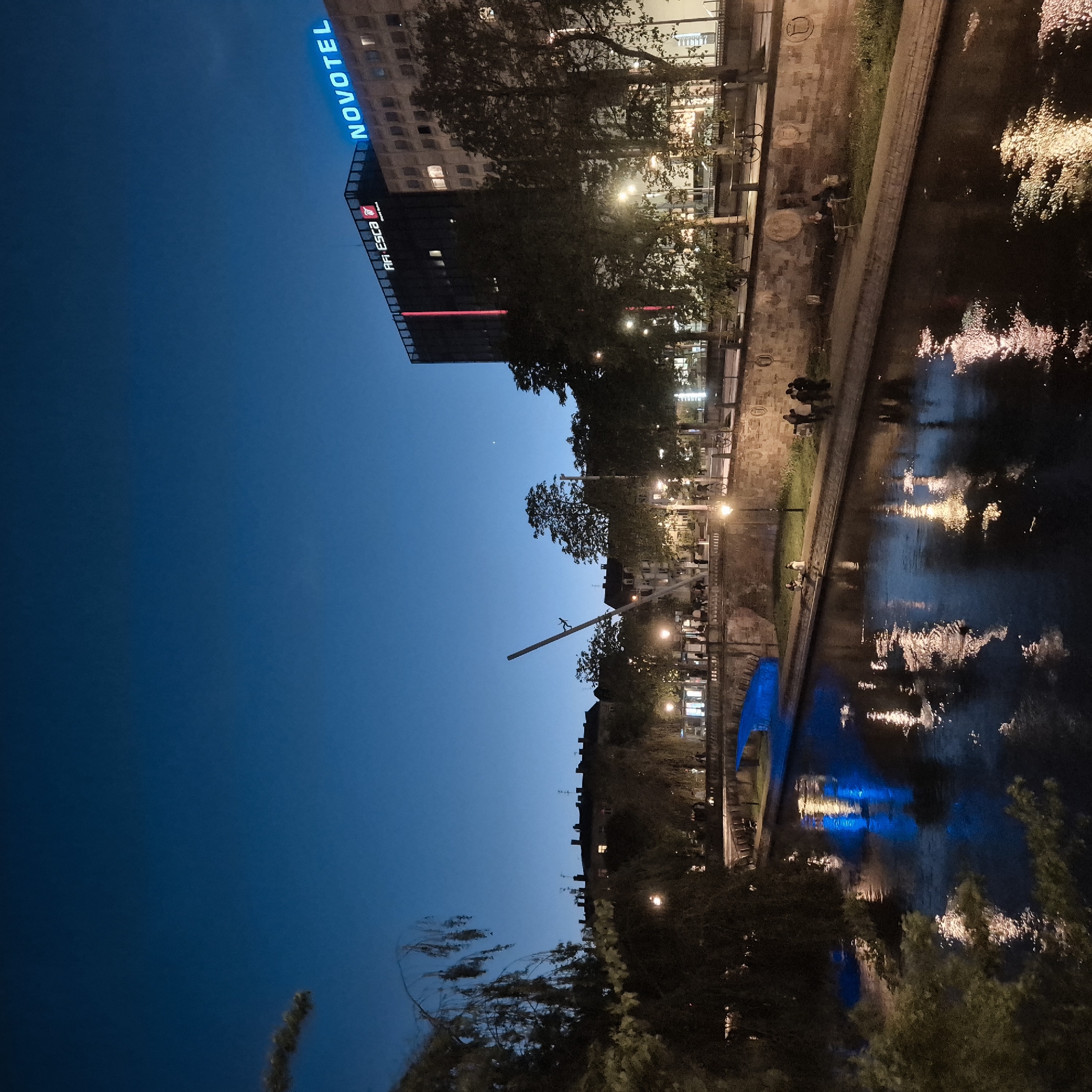 The image shows a nighttime urban scene in Strasbourg, France. A river runs along the right side of the image, reflecting lights. On the left bank is a stone embankment where people are sitting and standing. A building with the "Novotel" sign is visible on the upper left. The sky is a deep blue, transitioning to black at the bottom. Trees and streetlights line the riverbank.