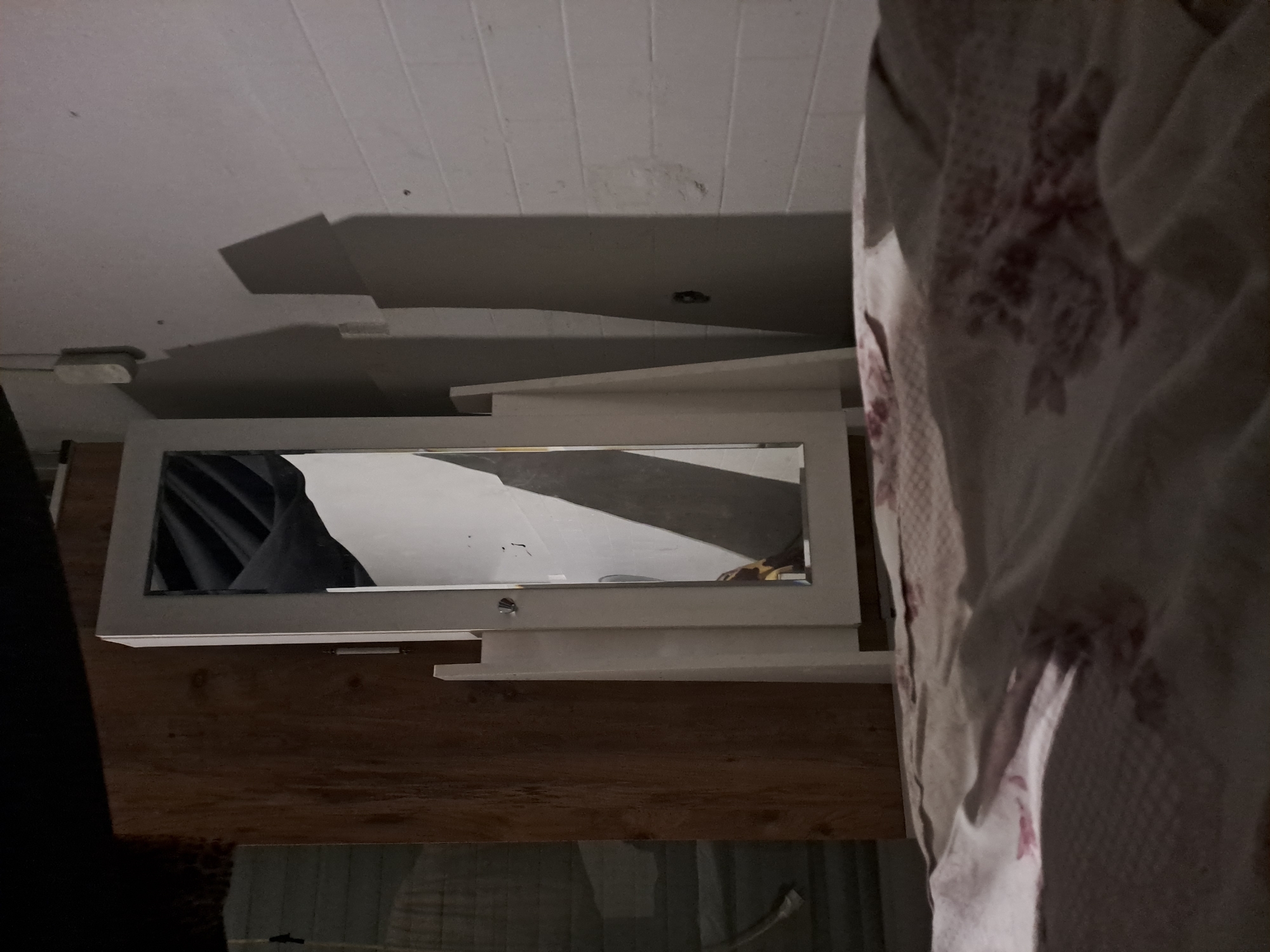 A full-length mirror on a stand is positioned next to a wooden cabinet. The mirror reflects a dark surface and a portion of a white wall. In the foreground, a crumpled bedsheet with a floral pattern is visible. The scene appears to be indoors, possibly in a bedroom. There is no indication of Manisa, Turkey.