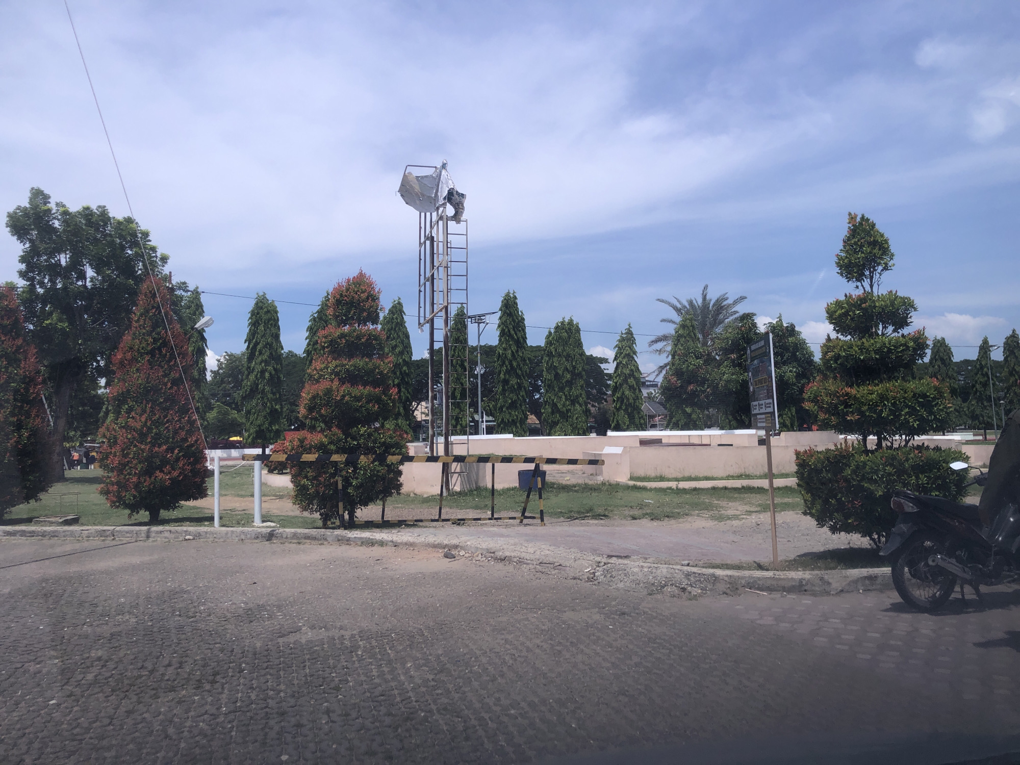 The image depicts a public space in Lhokseumawe, Indonesia, on a sunny day. The foreground shows a cobblestone road. A light-colored concrete wall with trees lines the background, creating a park-like setting. A metal structure with a large, partially covered reflector or light fixture stands out. An old-fashioned yellow and black parking barrier sits between trees in the center of the image. To the far right, partially obscured by a bush, is a parked black motorcycle. People are observed moving in the background.