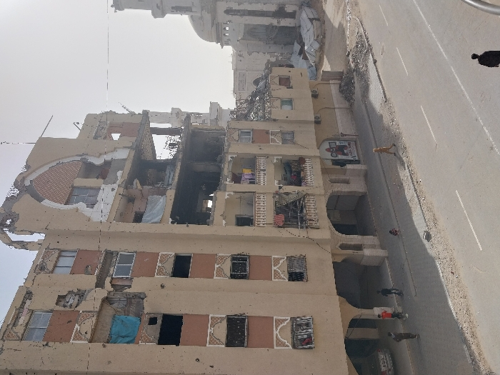 A severely damaged beige building stands next to a wide road. The building’s facade is missing in parts, revealing exposed brick and structural elements. Some windows are intact, while others are gaping holes. Laundry hangs from balconies. A dog and several people are seen on the road. A mosque with a satellite dish can be seen in the background. The sky is overcast and the image appears to depict a scene of war.