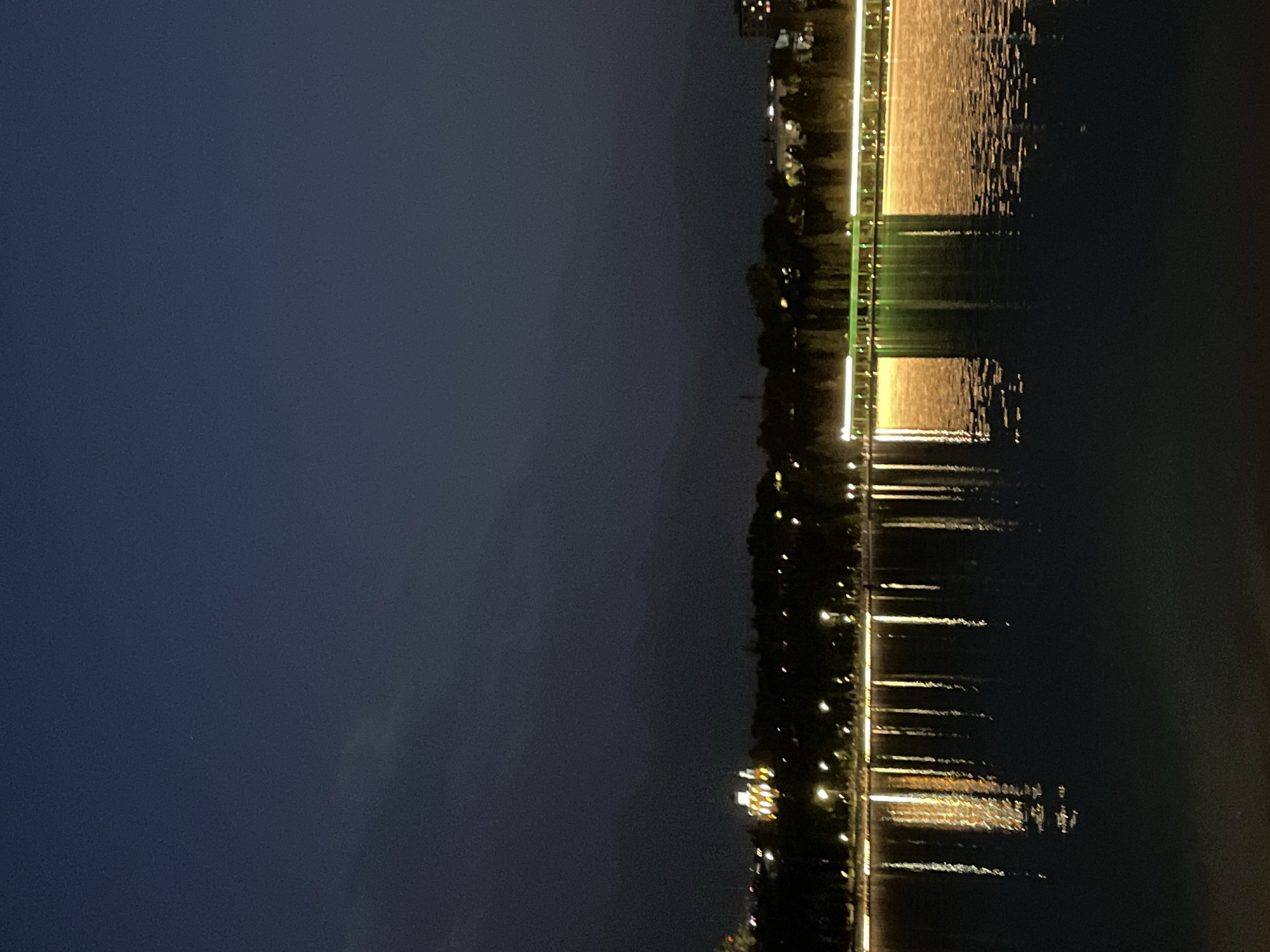 This image shows a nighttime scene at Kok Tobe Park in Almaty, Kazakhstan. The photograph is taken from across a body of water, likely a lake or pond, looking towards the park which is illuminated with numerous lights. The sky is dark blue, with some faint clouds visible. The lights from the park create long reflections on the water's surface, giving the impression of a shimmering, abstract painting. In the distance, a large, brightly lit building with a distinctive architecture stands out against the dark silhouette of trees. The overall atmosphere is serene and peaceful, capturing the beauty of the city at night.
