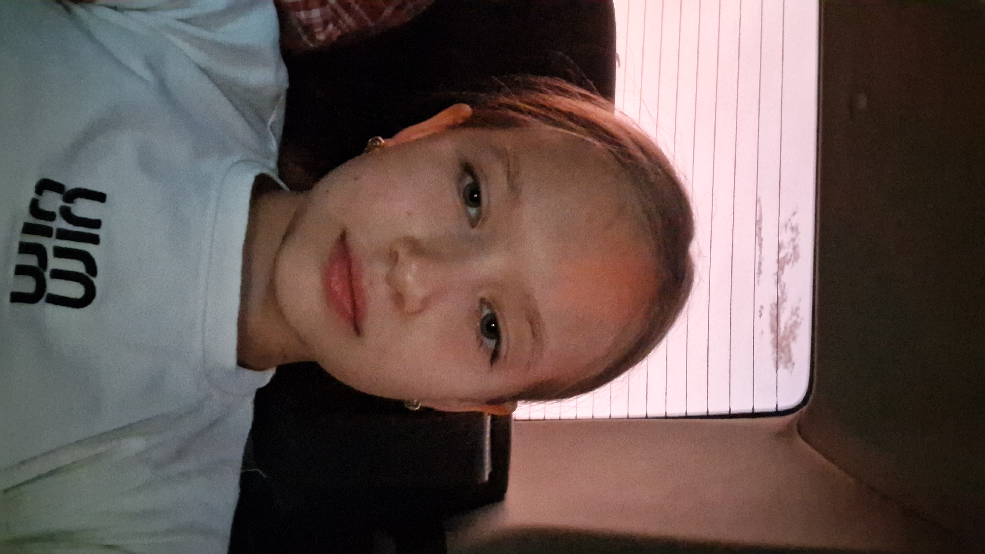A close-up, waist-up, selfie-style photograph captures a young Caucasian female with light brown hair pulled back, facing forward with a neutral expression. She is wearing a white t-shirt with a black graphic logo on the front. The background shows the interior of a vehicle, with a textured headrest visible behind her and a window with horizontal blinds in the upper portion. The lighting suggests it might be dusk or dawn, with a soft, warm glow illuminating the subject and the vehicle's interior. There are no specific geographical markers to indicate the city of Semey, Kazakhstan.