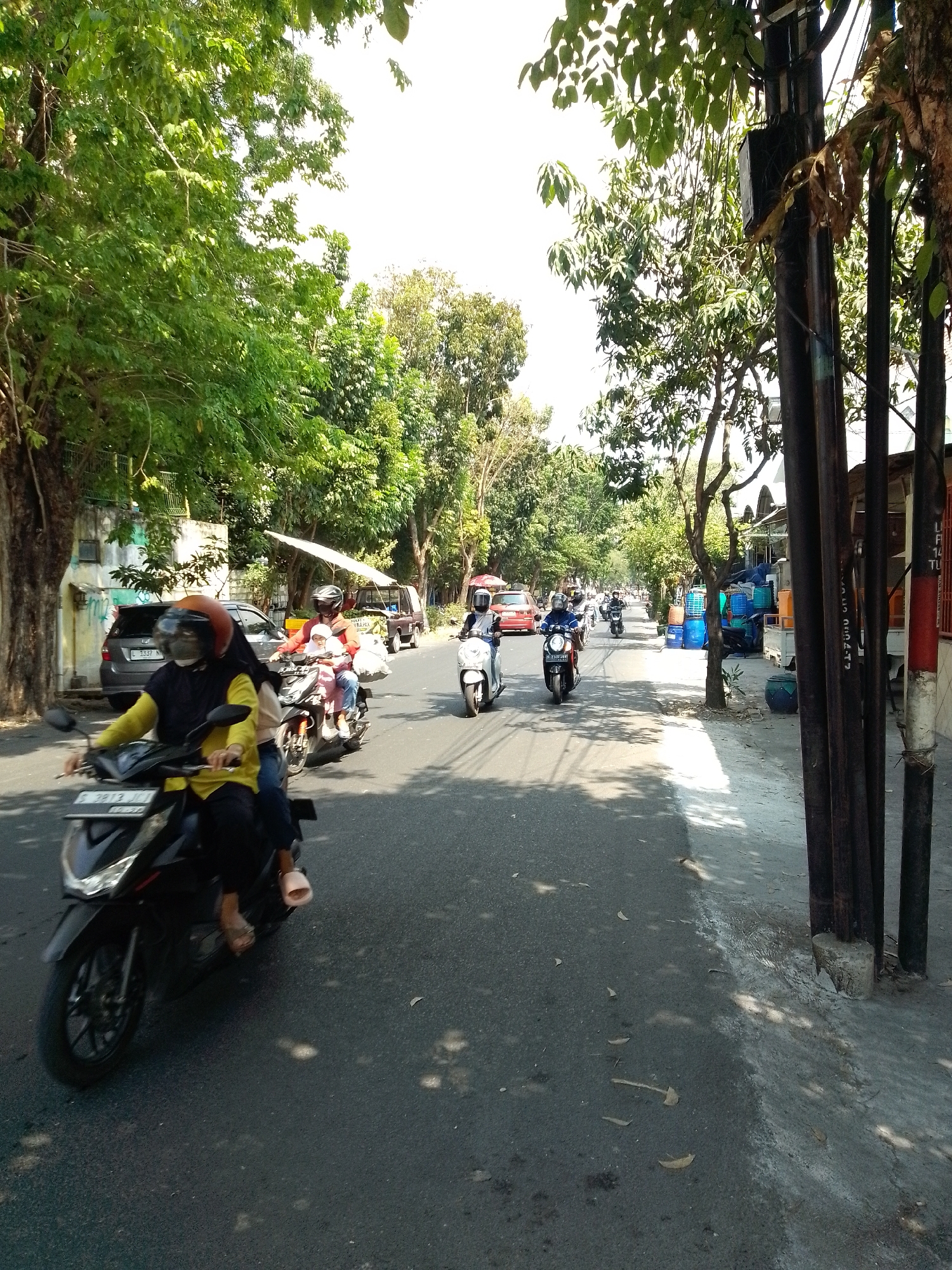 The image depicts a street scene in Tumenggungan, Indonesia, on a sunny day. The street is asphalt and appears to be moderately trafficked with several motorcycles and a few cars. On the left side of the road, there's a dark-colored car parked, and further down, a building with a green fence. Trees with lush green foliage line the left side of the street, casting shadows on the road. On the right side, there are several utility poles with wires, and further down, a street vendor with a white canopy selling goods, possibly including large blue plastic containers and smaller orange containers. People are seen riding on motorcycles, many wearing helmets and masks. One motorcycle in the foreground has two riders, one in a yellow shirt and black hijab, and another person behind them. Another motorcycle further ahead has a rider in a dark blue outfit with a white helmet, and a child passenger wearing a pink head covering. The overall atmosphere suggests a typical day in a moderately busy urban or suburban area.