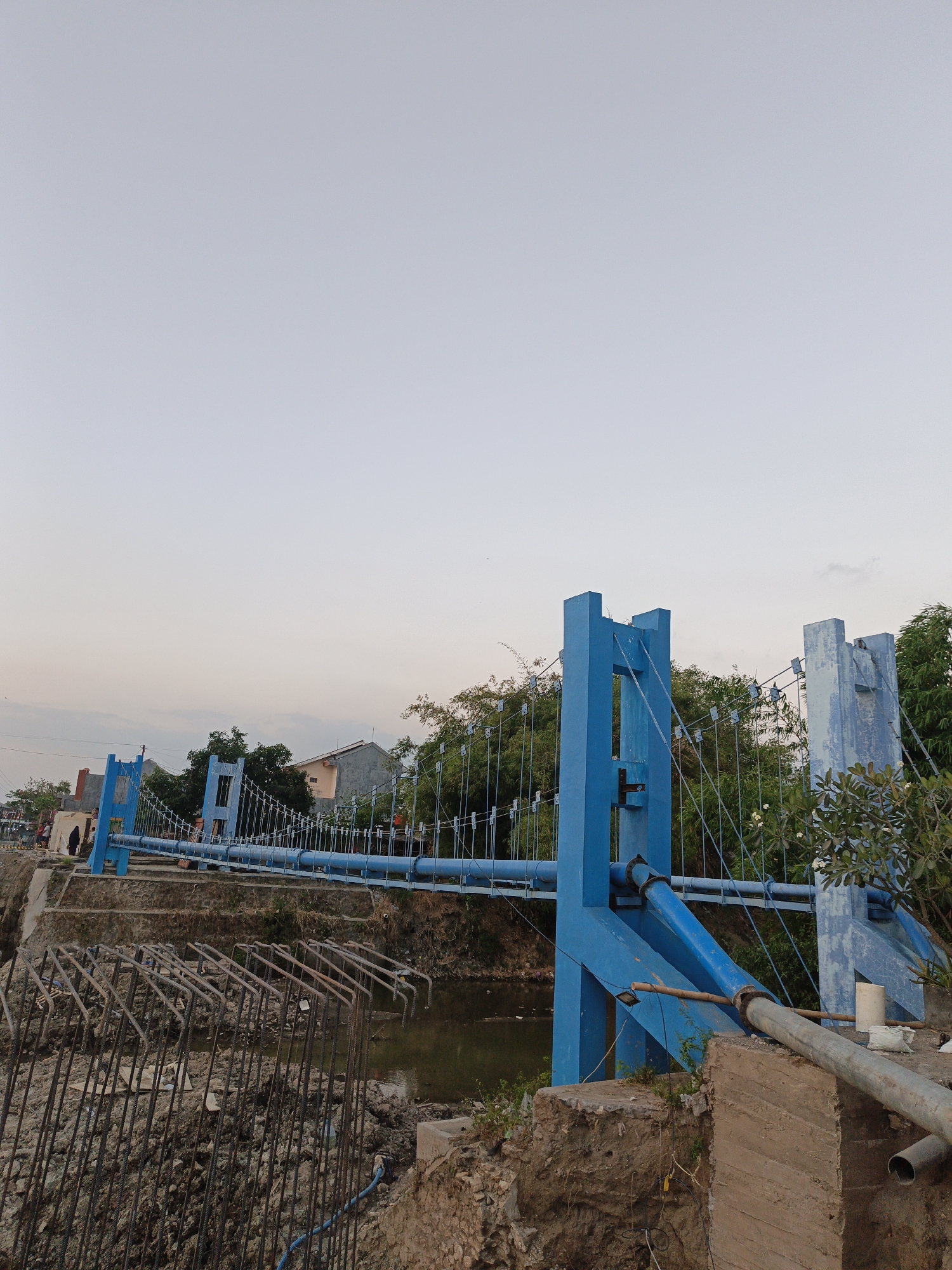 A suspension bridge with bright blue towers and railings spans a calm body of water. The bridge is suspended by wires attached to the towers. In the foreground, construction materials, including rebar and concrete blocks, are visible, suggesting ongoing or recent construction work on the riverbank. A dirt path leads up to the bridge on the left side, where a few people are seen walking. Trees and buildings line the opposite bank of the river. The sky is a pale, overcast gray, indicating early morning or late afternoon light. The scene appears to be in a semi-urban or rural area of Planggu, Indonesia, given the presence of residential buildings and a natural waterway.