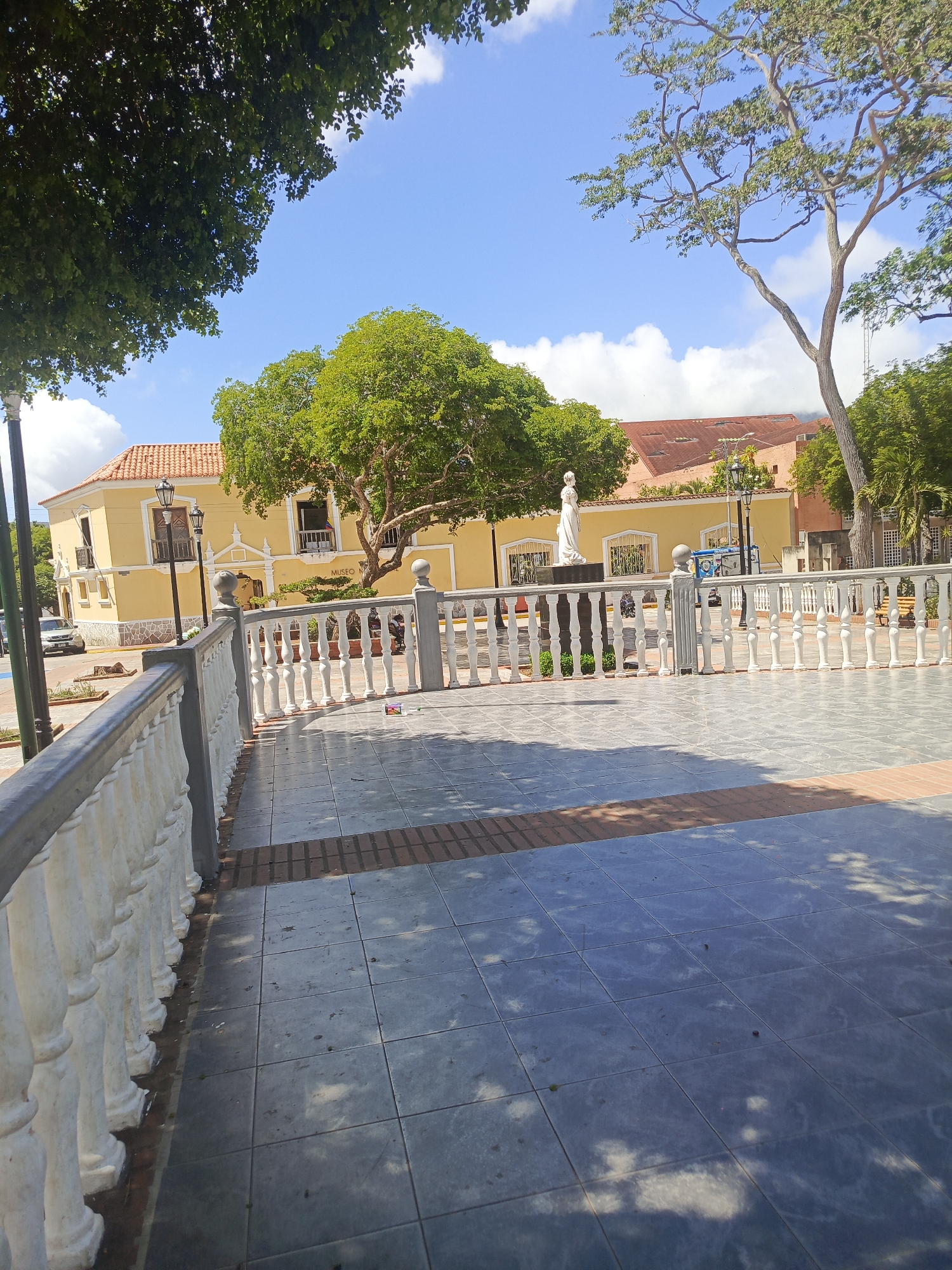 The image depicts a public square in La Asuncion, Venezuela. The foreground shows a tiled plaza with a low, white balustrade on the left and a similarly styled fence in the middle ground. Behind the middle ground fence stands a white statue, possibly a monument or figure. The background features yellow colonial-style buildings with red tile roofs and trees. The sky is blue with some clouds. No people are visible in the scene.