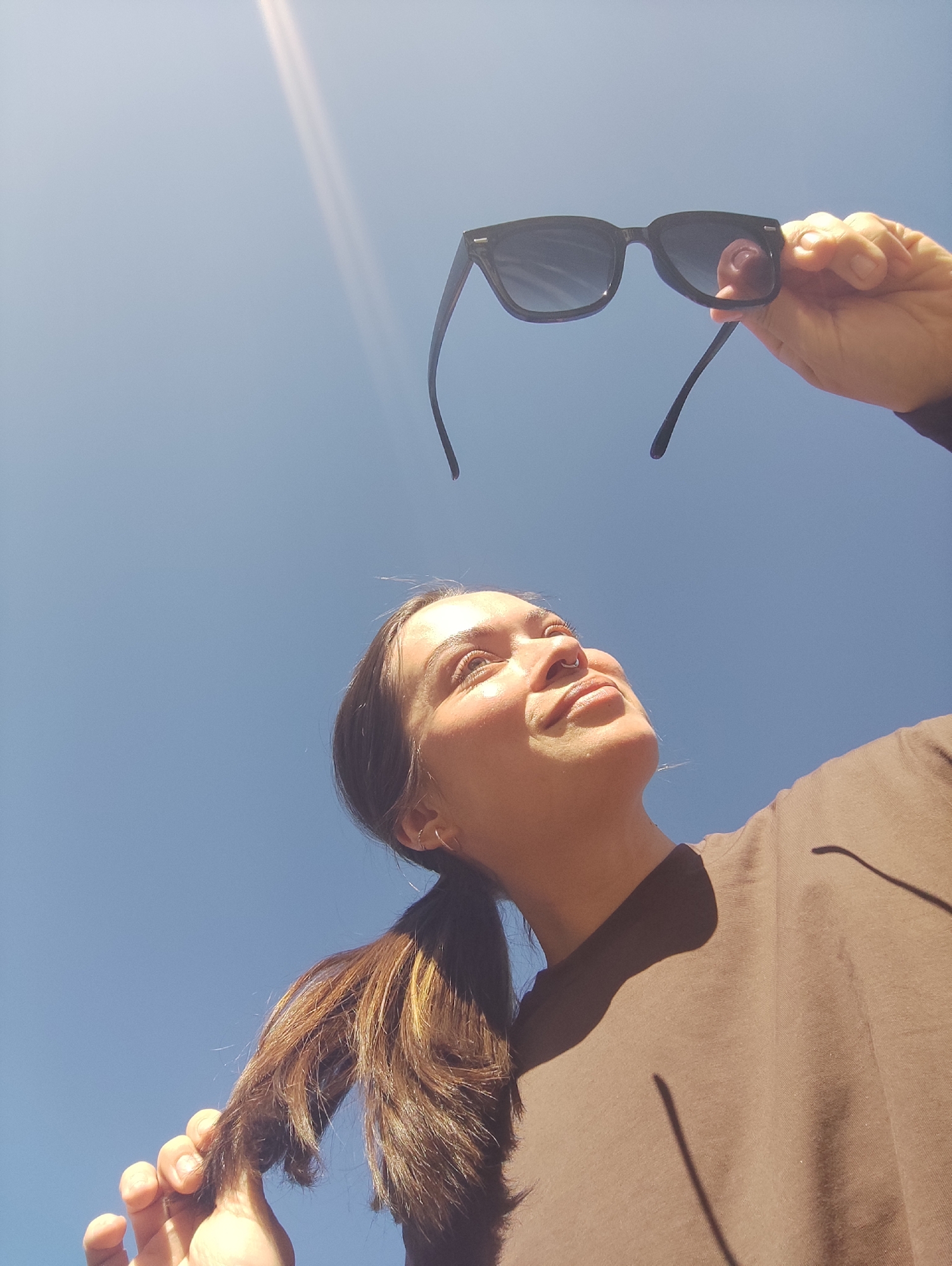 A woman stands under a bright, clear, blue sky. She is holding a pair of black sunglasses aloft, examining them or shielding her eyes. She has brown hair pulled back into a low ponytail and wears a brown shirt. The sun is shining directly above, creating a bright streak in the sky. The photograph appears to be taken from a low angle, looking up towards the woman and the sky. The location in the city of Фоки, Russia is not discernible from the image.
