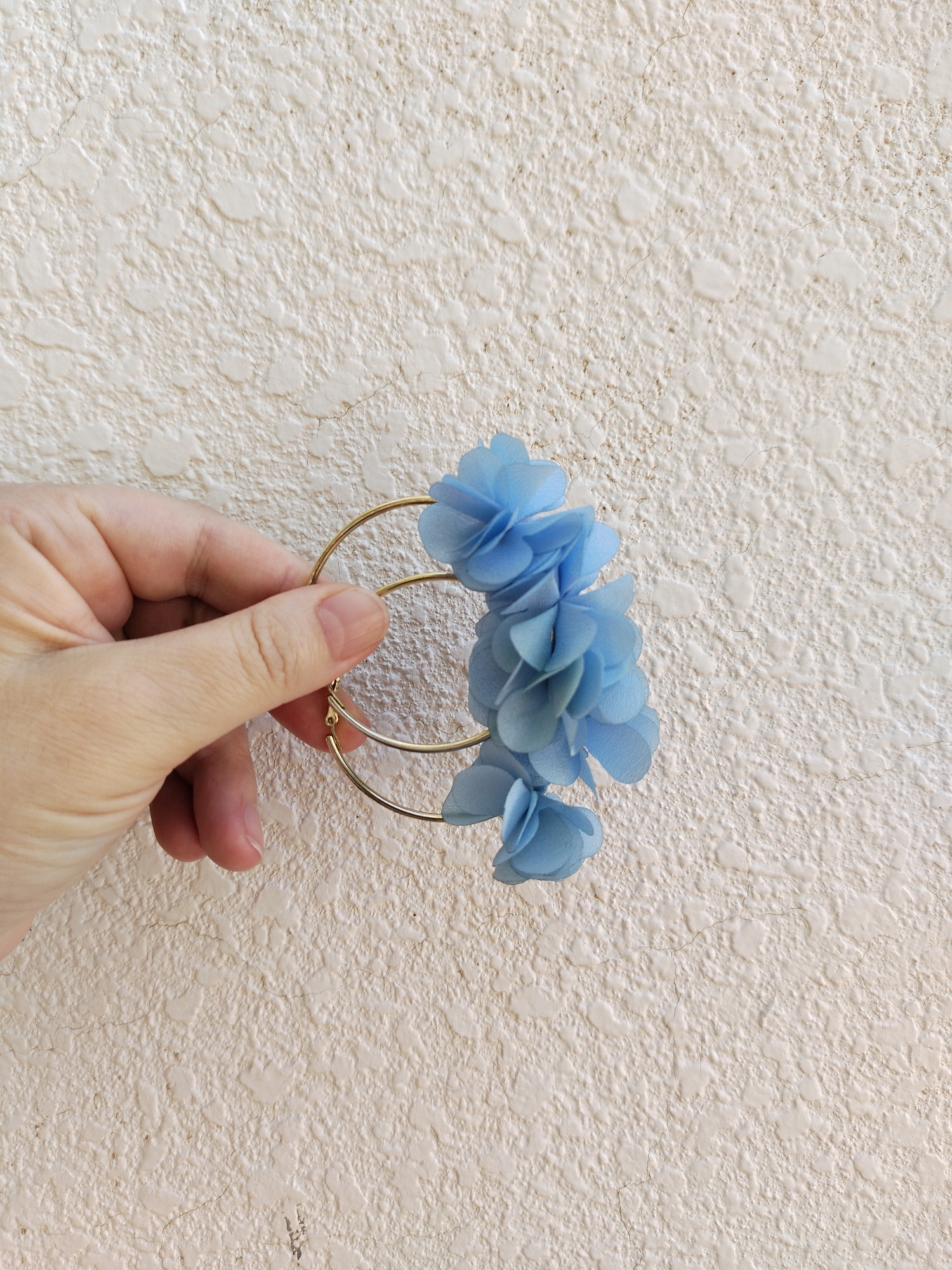A hand is holding three golden hoop earrings, each adorned with several light blue, fabric flowers. The background is a textured, light-colored wall. The location of the image is unknown.