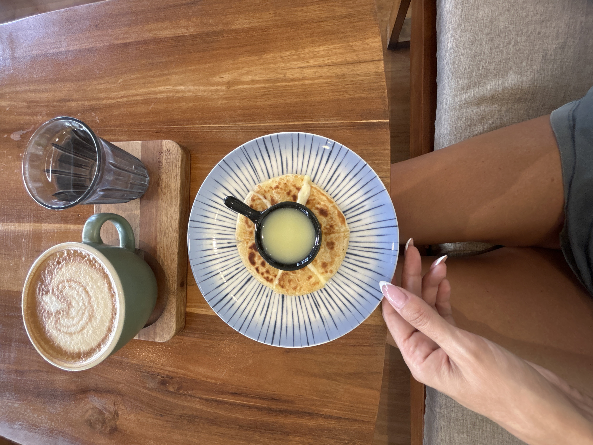 A overhead shot shows a person's hands and legs sitting at a wooden table in a cafe in Mengwi, Indonesia. On the table, there is a green mug with latte art, a glass of water, and a plate with a flatbread and a small bowl of dipping sauce. The person's left hand, with french manicured nails, is reaching towards the plate. The background consists of the wooden table and the person's legs, covered by a gray blanket and a dark gray shirt. The cafe appears to be outdoors or has an open-air concept due to the natural lighting.