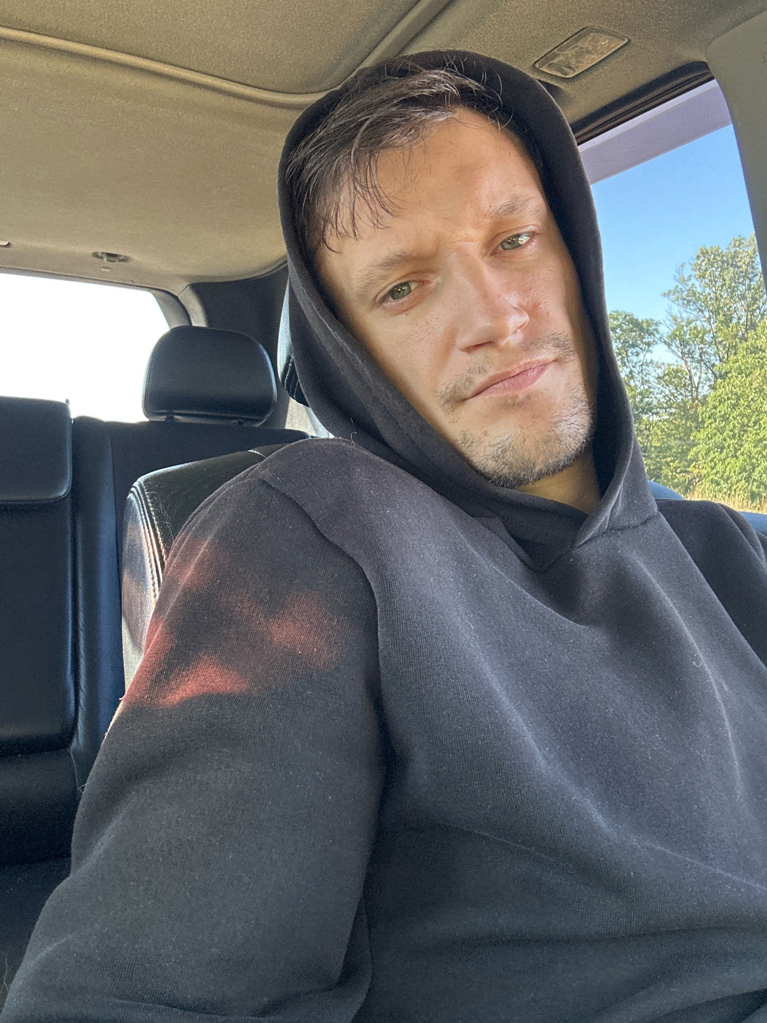 The image is a selfie taken by a man in the backseat of a car. He is wearing a black hoodie and has a beard. The interior of the car is visible, including black leather seats and the car's ceiling. Outside the car, trees and a clear blue sky are visible. The image does not provide any specific information about the location within the city of Makiv, Ukraine, or any of its landmarks.
