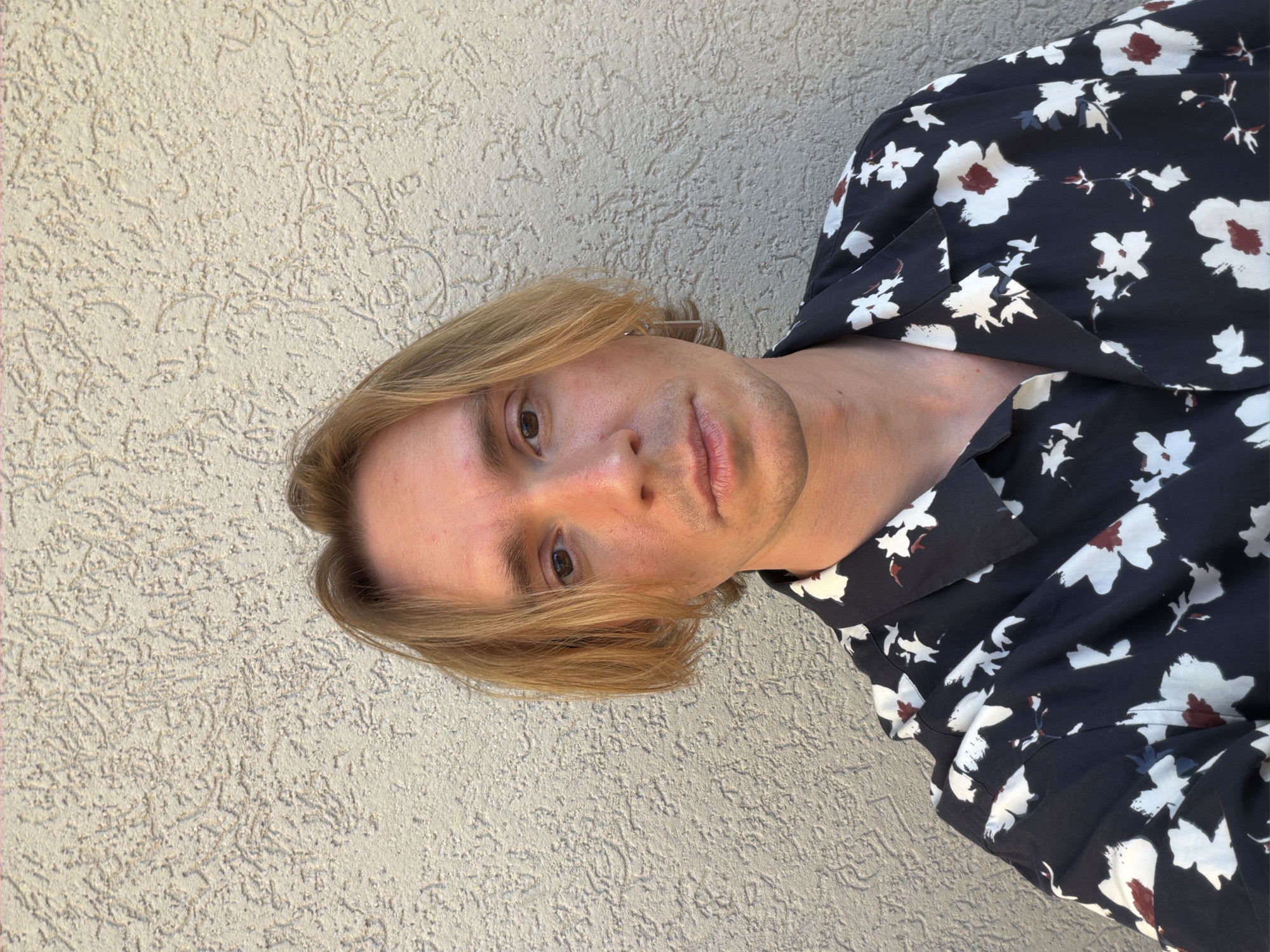 A medium close-up, portrait-style photograph captures a person, likely male, with light blonde, medium-length hair, looking directly at the camera with a neutral expression. The image is rotated 90 degrees clockwise, placing the subject's head towards the left side of the frame and his body extending downwards. He has fair skin, visible stubble on his chin and jawline, and brown eyes. He is wearing a dark (possibly navy blue or black) collared shirt adorned with a floral pattern of white petals and reddish-brown centers.
The background consists of a uniformly textured, light beige or off-white stucco wall, suggesting an outdoor setting. The lighting is bright and natural, indicating daytime, with direct sunlight casting soft shadows and highlighting the subject's features and hair. No specific activity beyond posing for the photograph is visible. There is no visible text in the image. The location context provided is Budva, Montenegro.