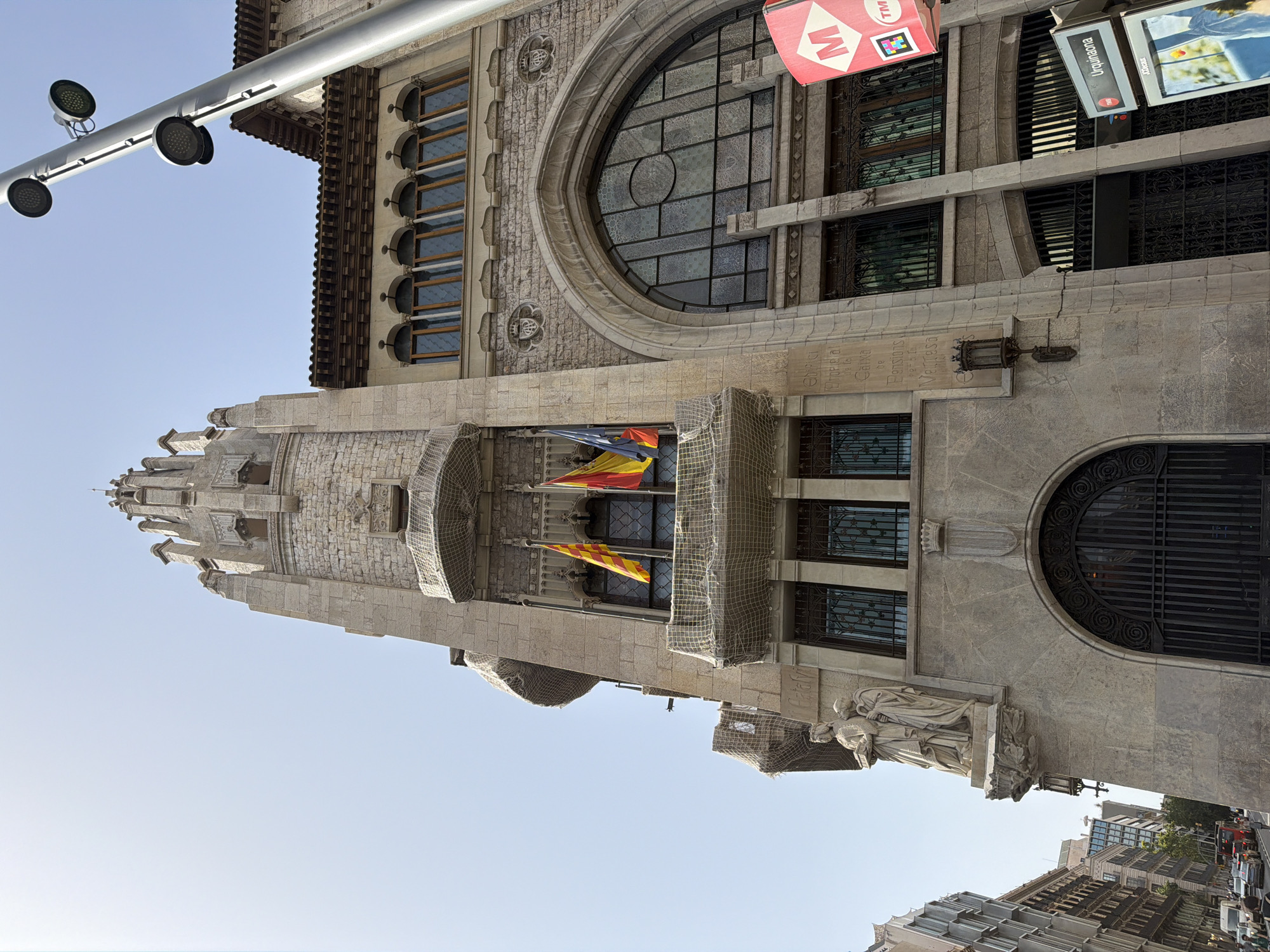 The image shows the exterior of the Edifici del Sindicat de la Industria Metal·lúrgica, a building located in Barcelona, Spain. The architecture features a prominent tower with Gothic-inspired detailing, stone carvings, and multiple balconies. Catalan flags are visible flying from the tower. The building's facade is made of stone and includes arched windows with intricate metalwork and stained glass. A large arched entrance with decorative metal gates is present. A statue of two figures stands on a projecting ledge on the left side of the building. Street level activity includes vehicles and pedestrians, with a "Urquinaona" metro station sign visible to the right of the building. The sky is clear and blue, suggesting a sunny day.