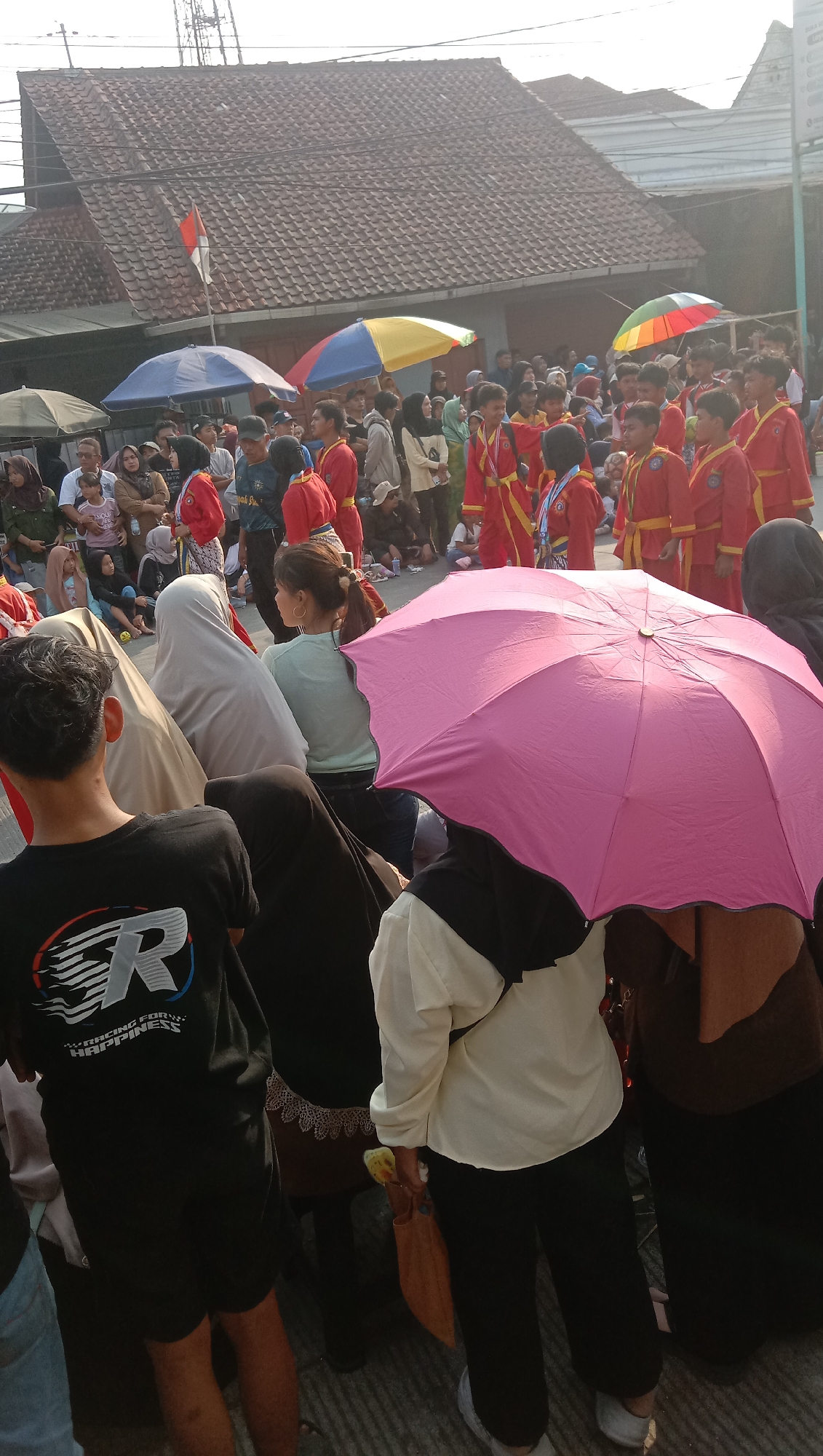 In Jatisawit, Indonesia, a crowd gathers outdoors, seemingly for a parade or procession. A group of young people in matching red and yellow martial arts uniforms stand in formation, some wearing medals. Spectators, many wearing headscarves, are positioned behind them. Colorful umbrellas, including a prominent pink one, provide shade. A building with a tiled roof and a red-and-white Indonesian flag is visible in the background. The ground appears to be paved.