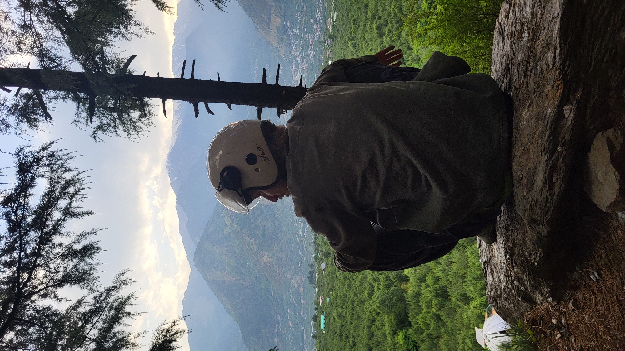 A person wearing a white helmet and dark clothing is sitting on a rocky ledge overlooking a scenic view. In the background is a lush green valley with distant mountains and a hazy sky. Tree branches frame the left side of the image, adding depth to the scene. The location is likely in the city of Nashala, India, judging from the terrain and vegetation.